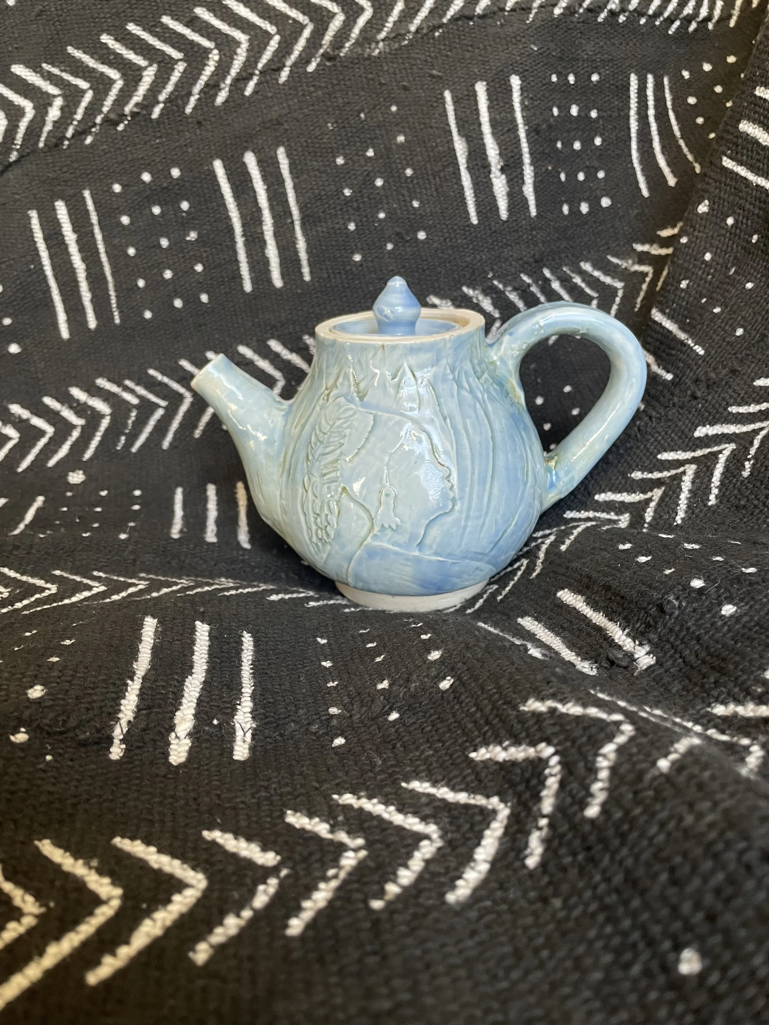 A small ceramic teapot with a light blue glaze, sitting on a black fabric with a white geometric pattern.