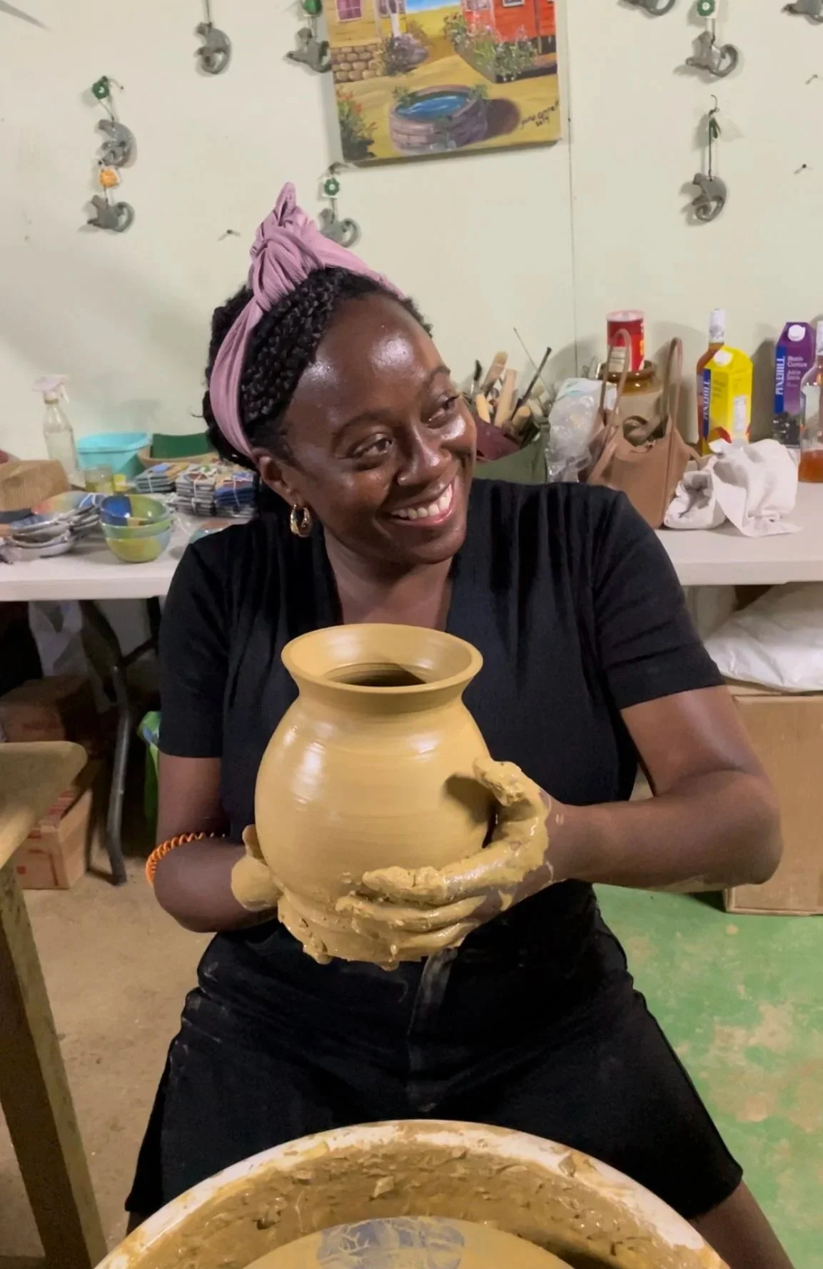 A woman with a pink headwrap and earrings is smiling while holding a large, partially crafted pottery vase in a ceramics studio. The background shows pottery supplies, brushes, and a colorful painting on the wall.