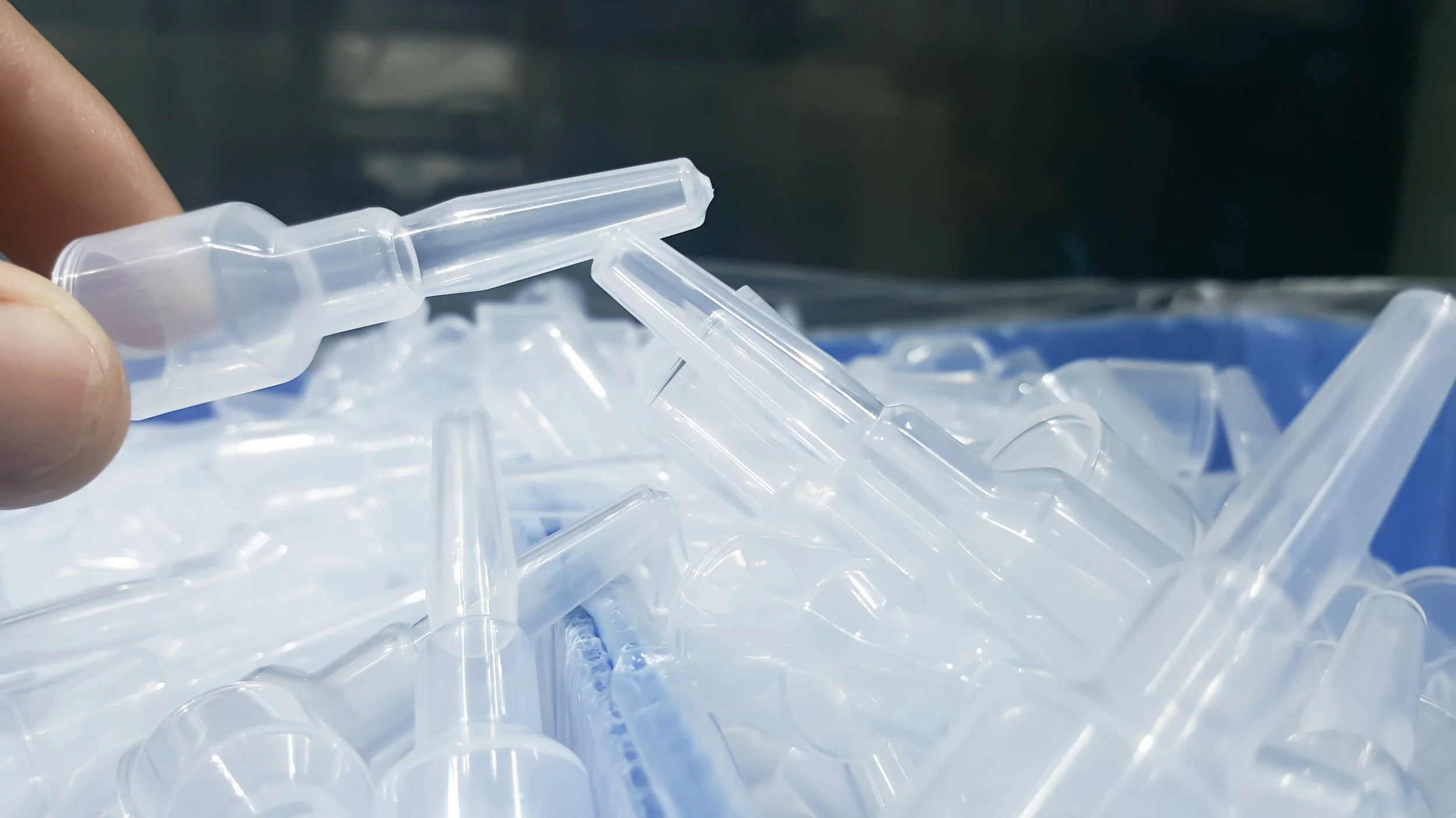 Close-up of a person holding a clear plastic medical tube, with more similar tubes scattered in a blue container in the background.