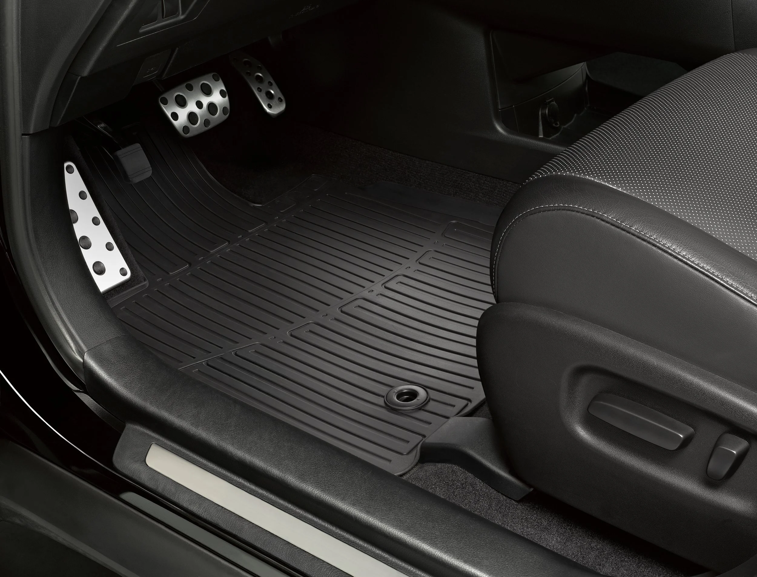Close-up of the driver side footwell area of a car, showing the accelerator, brake, and clutch pedals with metallic finishes and rubber grips, along with a black rubber floor mat.