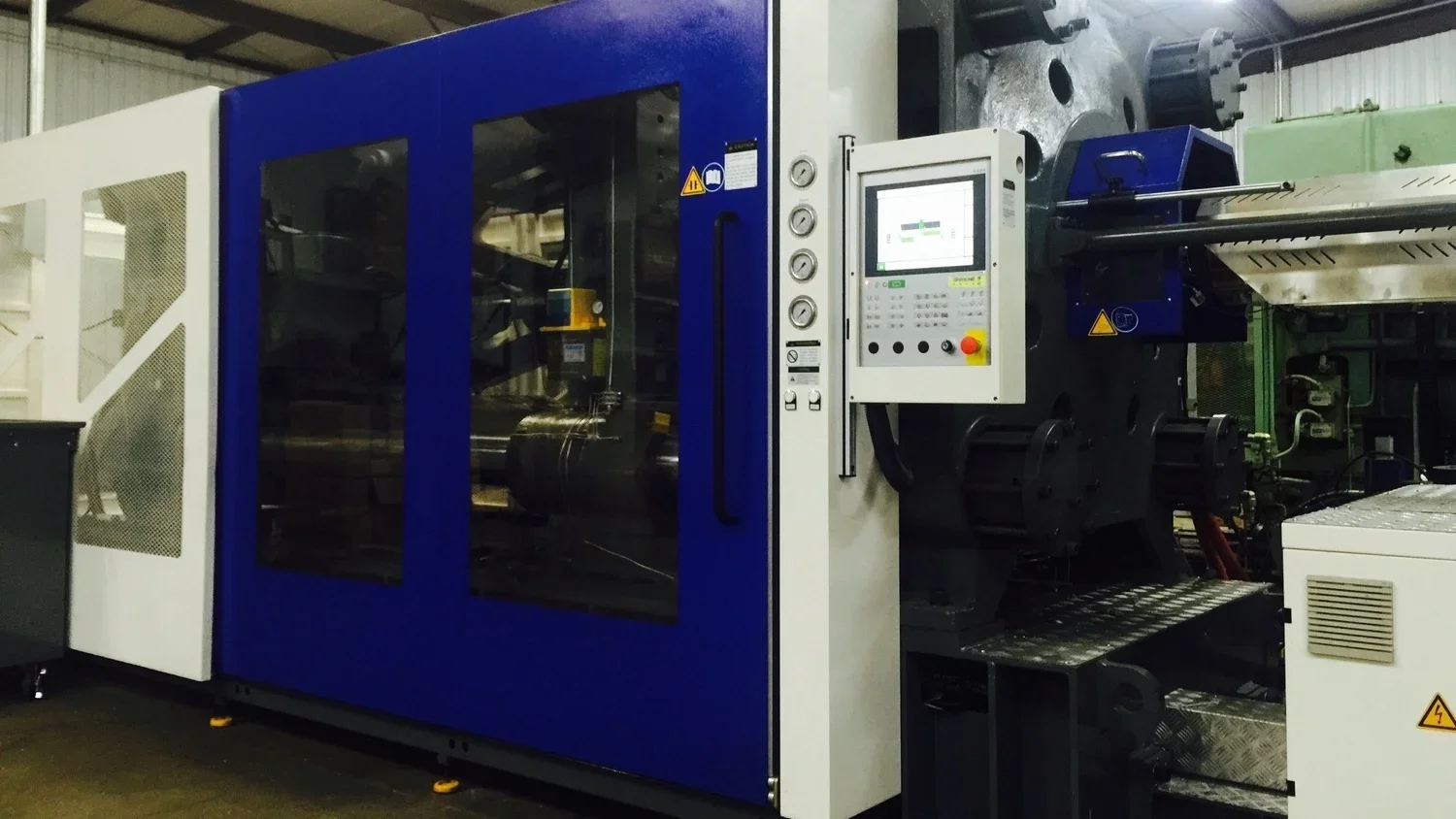 Large industrial machine with blue and white panels, a control panel with a screen and buttons, and metal rollers, inside a manufacturing facility.