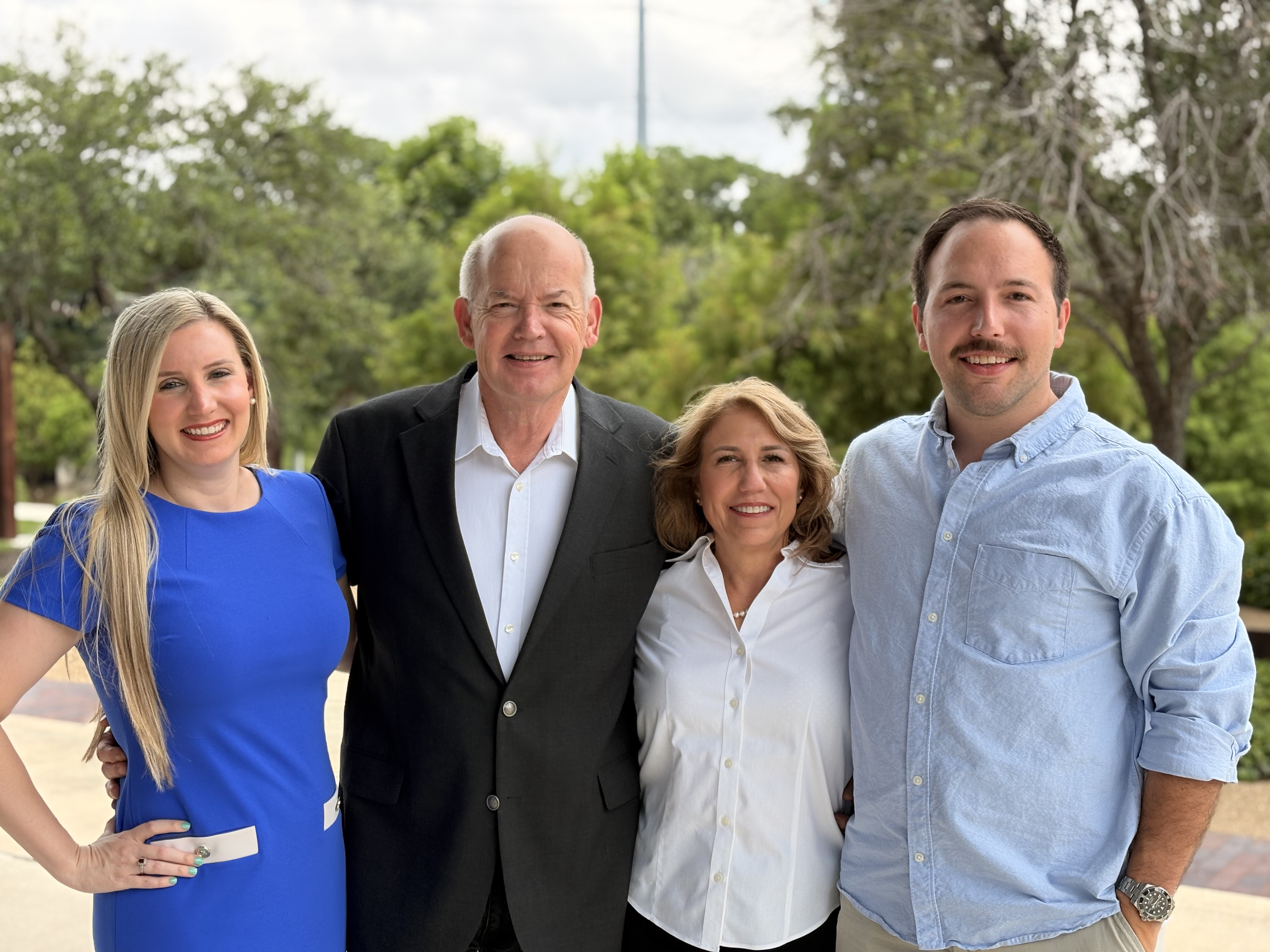 Group of five people standing outdoors with trees in the background, smiling.