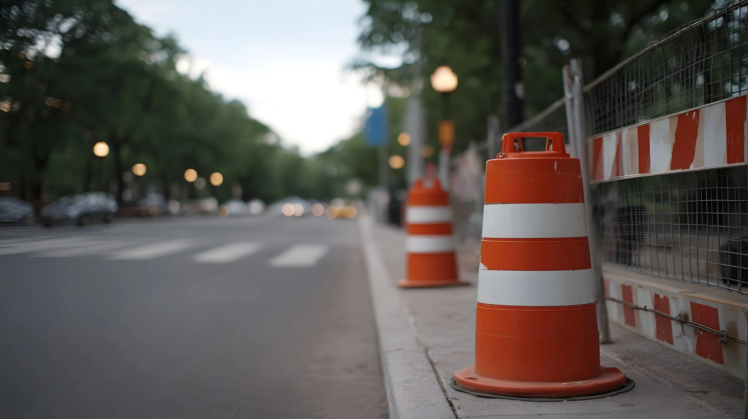Orange and white construction cones placed along a city sidewalk, with a construction barrier on the right, and trees and blurred street scene in the background.