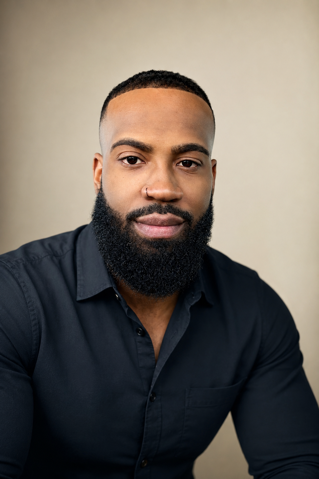 A portrait of a young man with a well-groomed beard and short hair, wearing a black button-up shirt, against a neutral plain background.