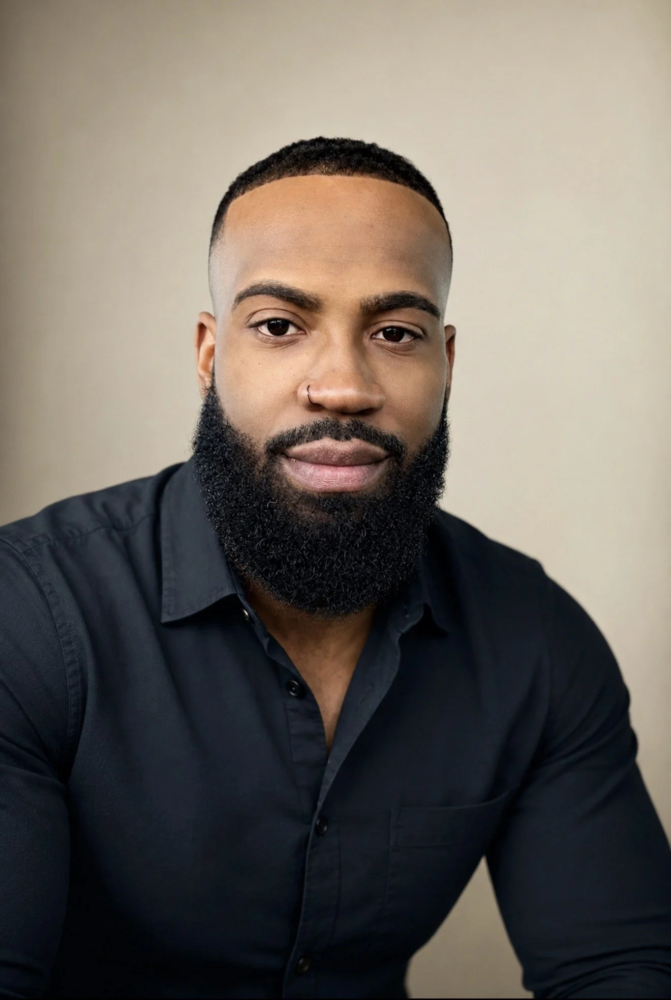 Close-up of a man with a well-groomed beard, short hair, wearing a black button-up shirt, against a plain beige background.