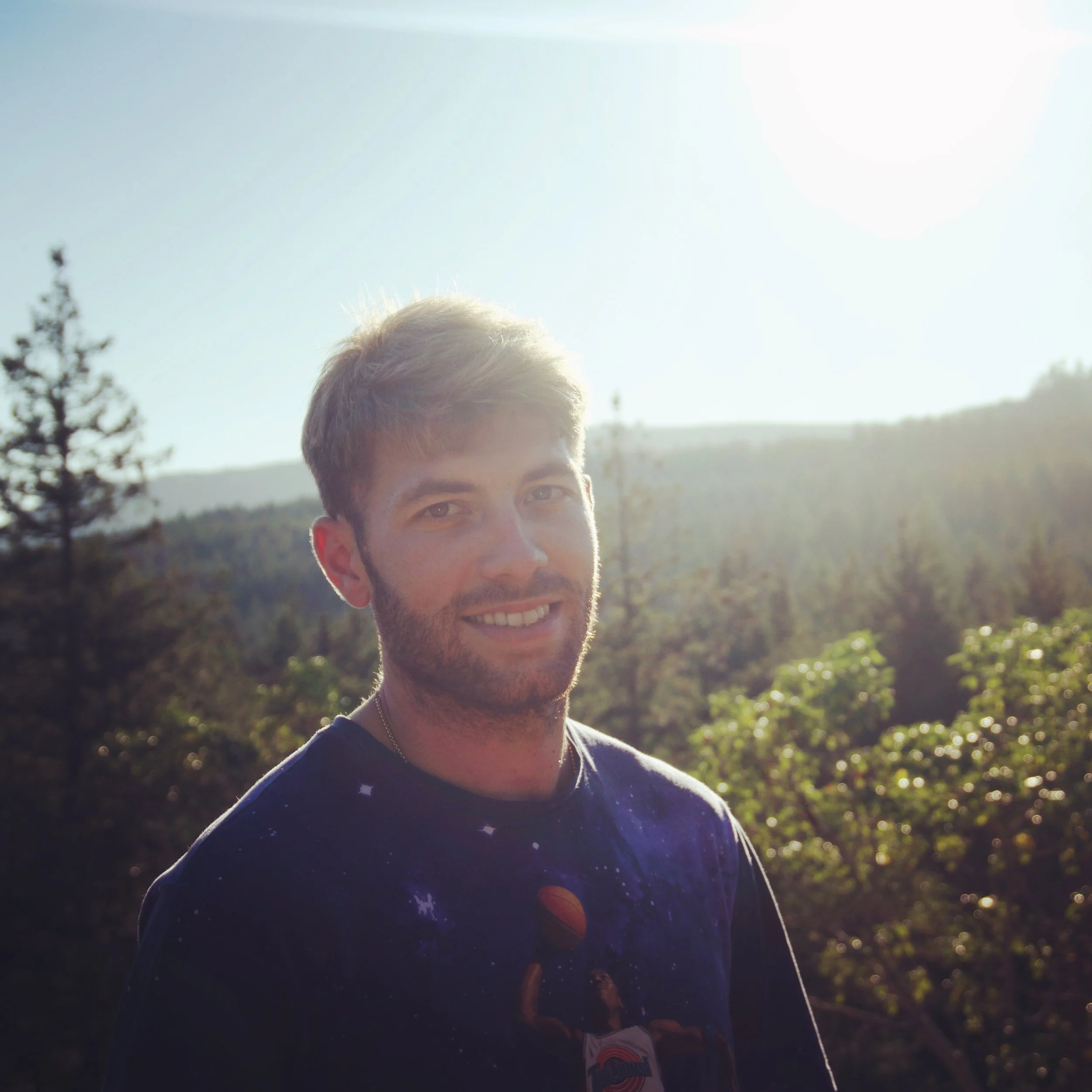 A person with short hair and light skin smiling outdoors with sunny weather, trees, and mountains in the background.