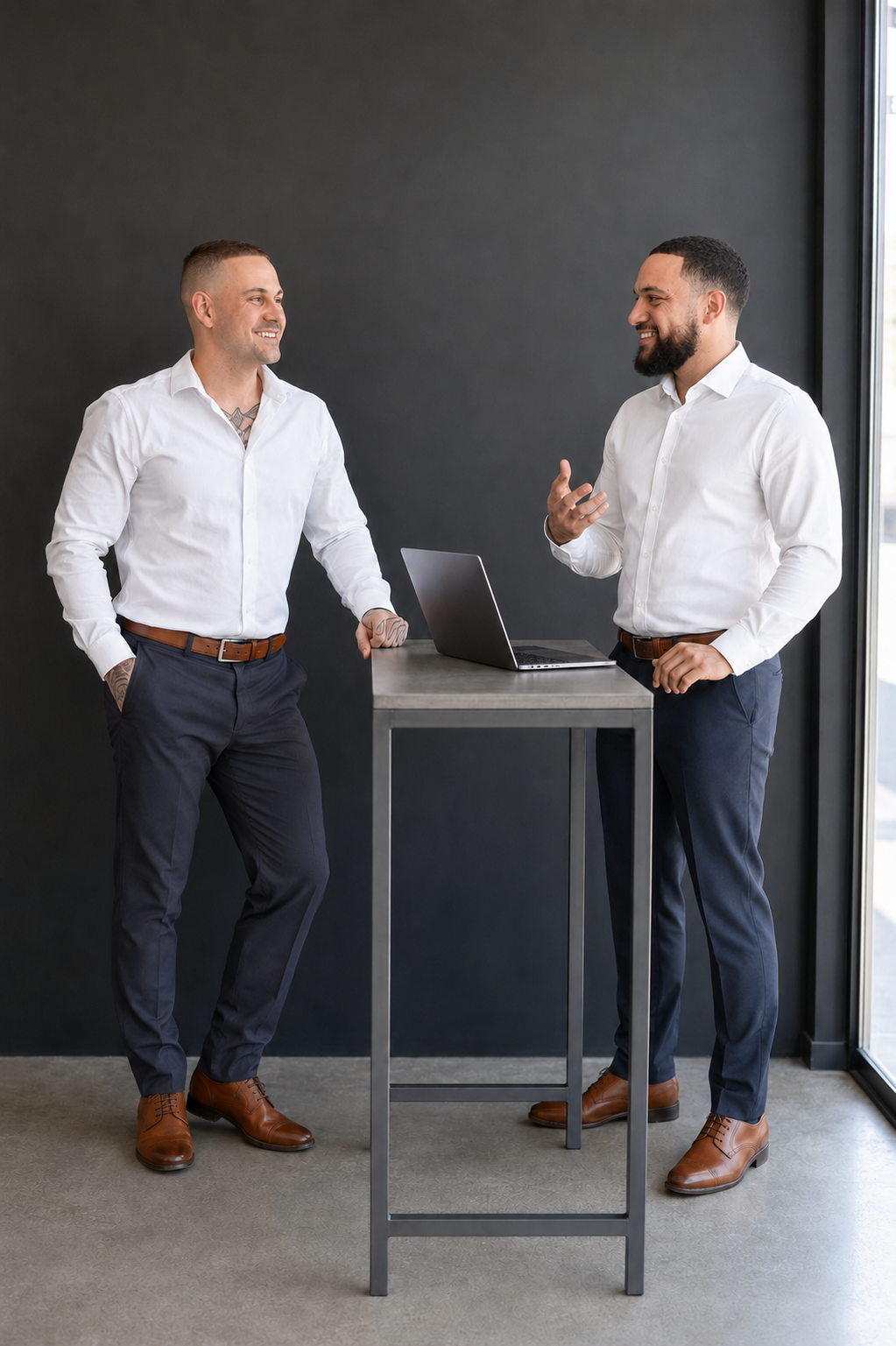 Two men in business attire having a conversation near a laptop on a high table in a modern office.