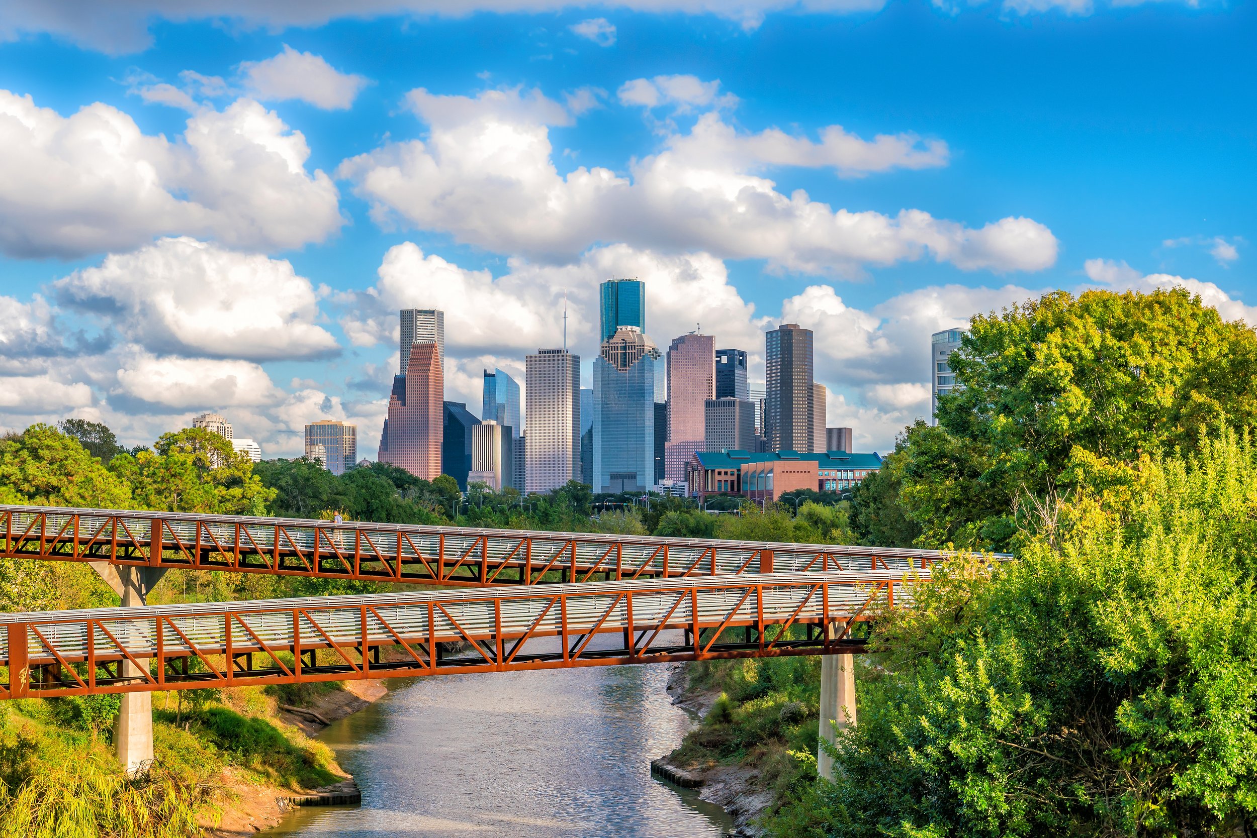 Houston City skyline with tall skyscrapers, a blue sky with clouds, and a river in the foreground. There are green trees and a bridge over the river.