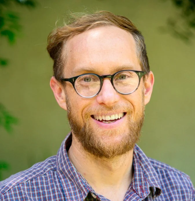 A smiling man with glasses, a beard, and light brown hair, wearing a checkered shirt with a green background.
