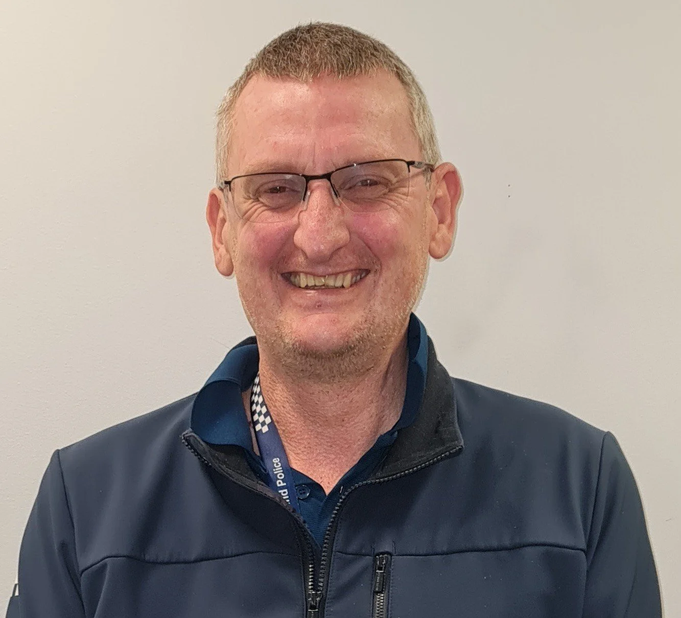 A man with glasses and short hair smiling, wearing a dark jacket and a police lanyard, standing against a plain light-colored wall.