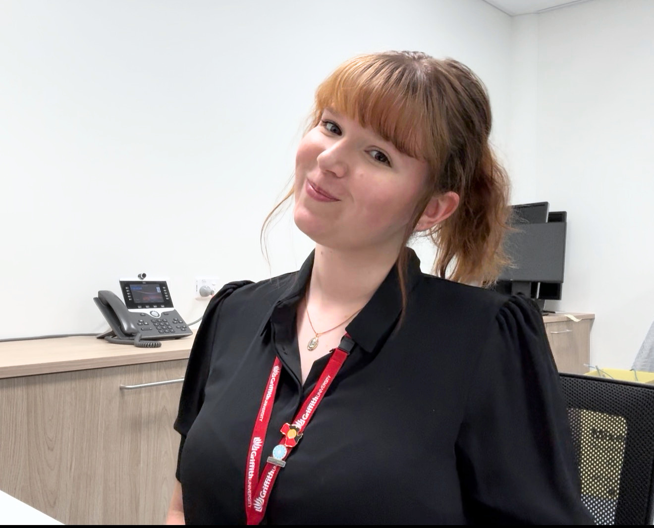 A young woman with reddish-brown hair in a ponytail, wearing a black blouse and a red lanyard, sitting in an office smiling.