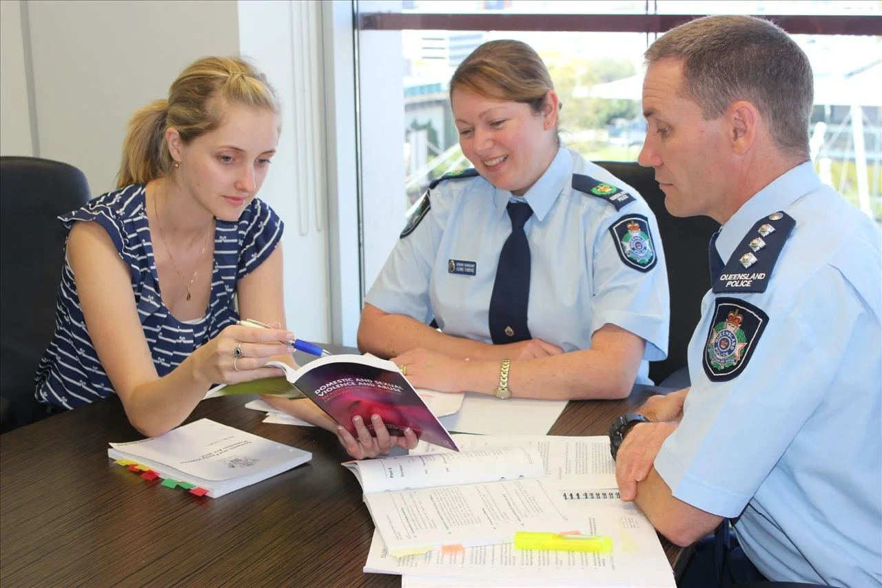 A woman sitting at a desk with two police officers, reviewing documents and a booklet about domestic and family violence, in an office with large windows.