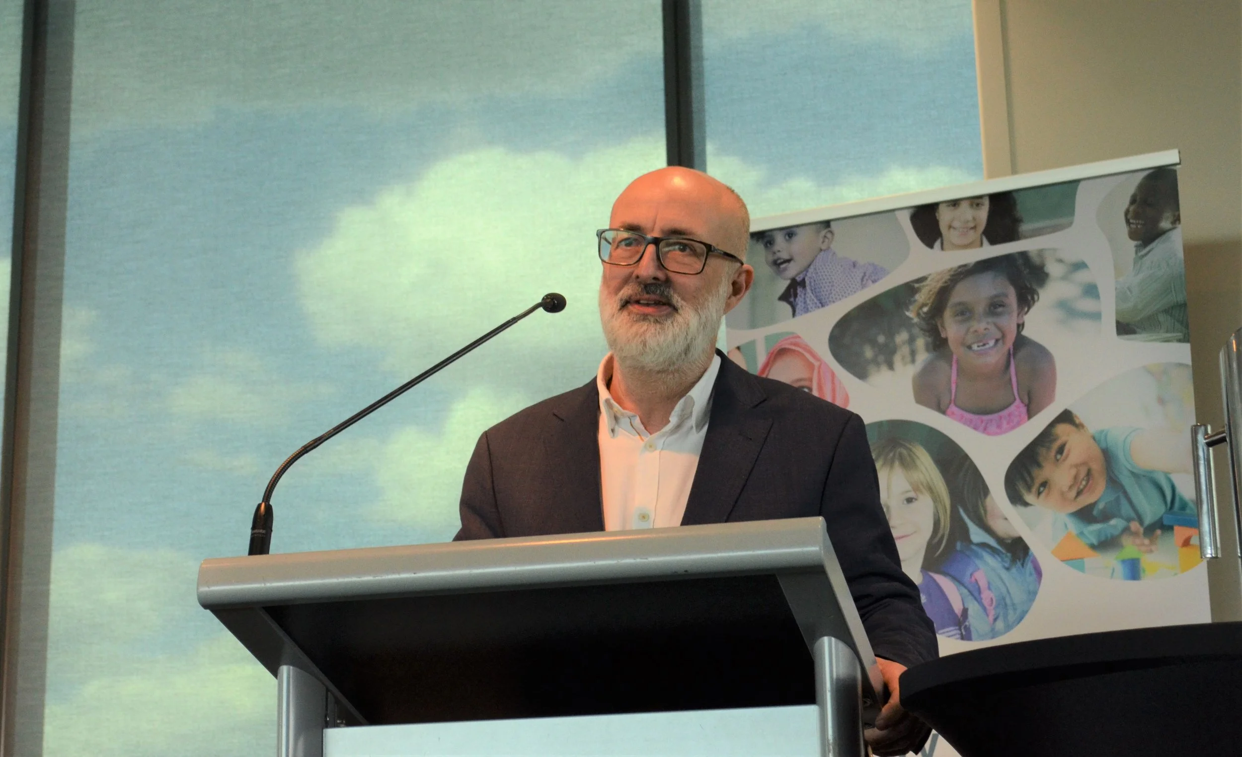 A man with glasses and a beard wearing a dark suit and white shirt standing at a podium, speaking into a microphone, with a large window and a banner with smiling children in the background.