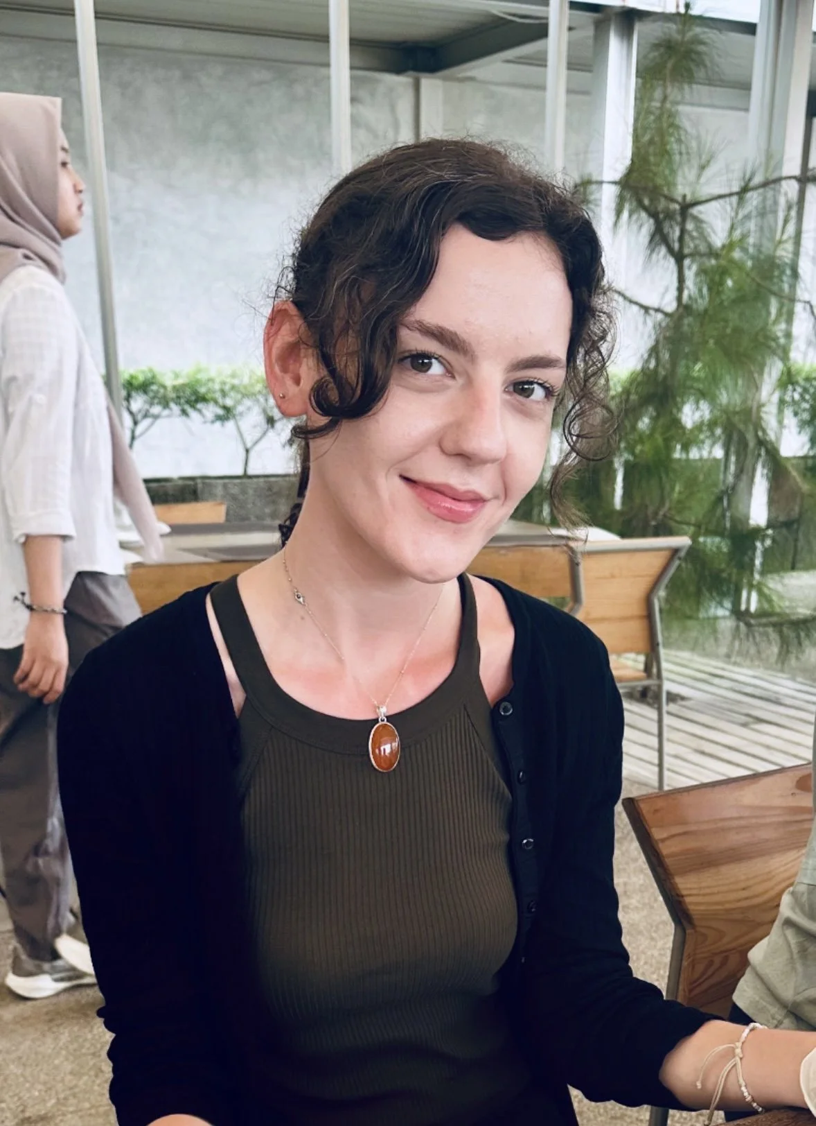 Close-up of a young woman with short, curly brown hair, wearing a dark green top, black cardigan, and a necklace with a large amber pendant, smiling at the camera in a bright indoor space with plants.