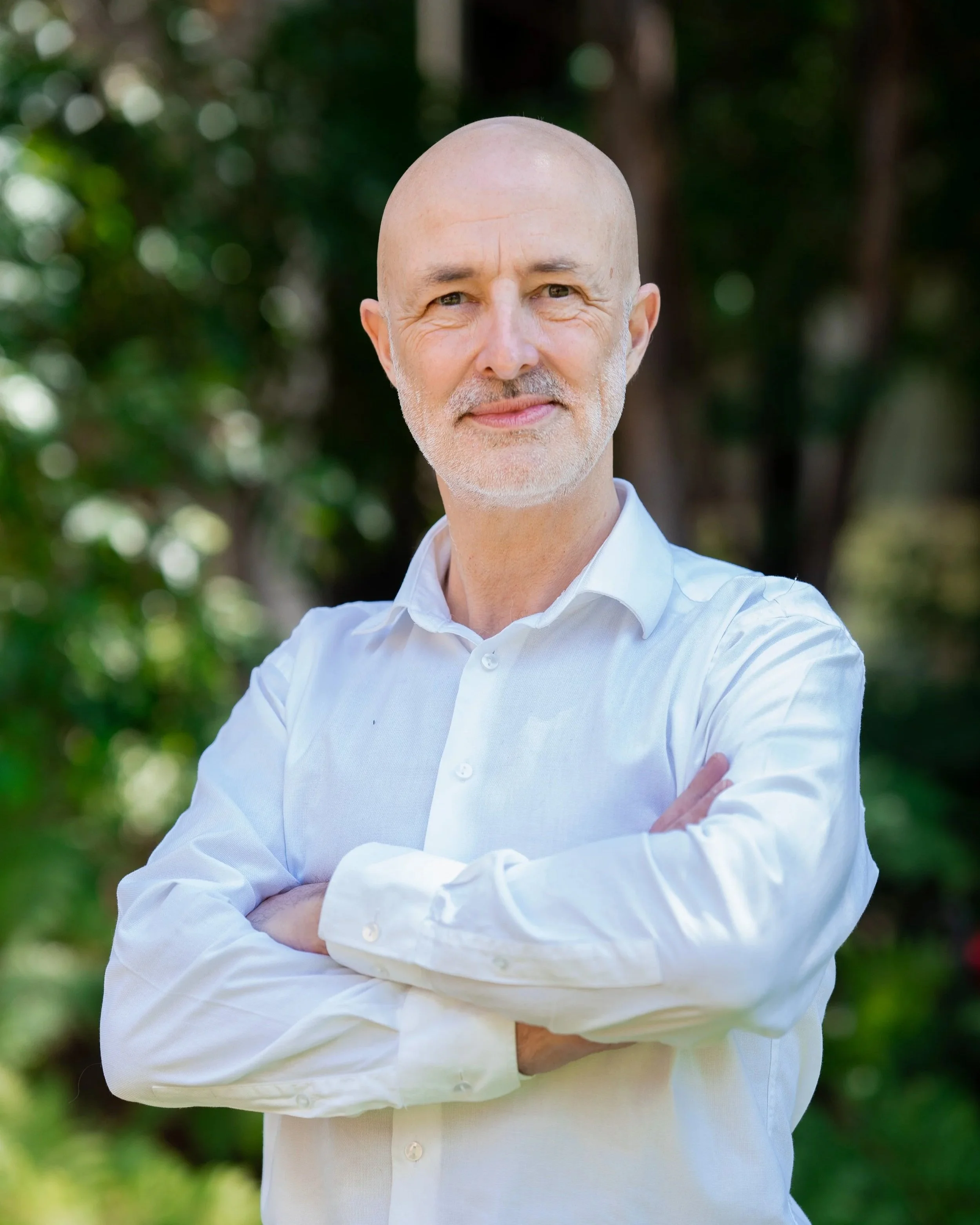 Close-up of a bald, middle-aged man with a beard and mustache wearing a white shirt, standing outdoors with crossed arms, smiling, with blurred green trees in the background.
