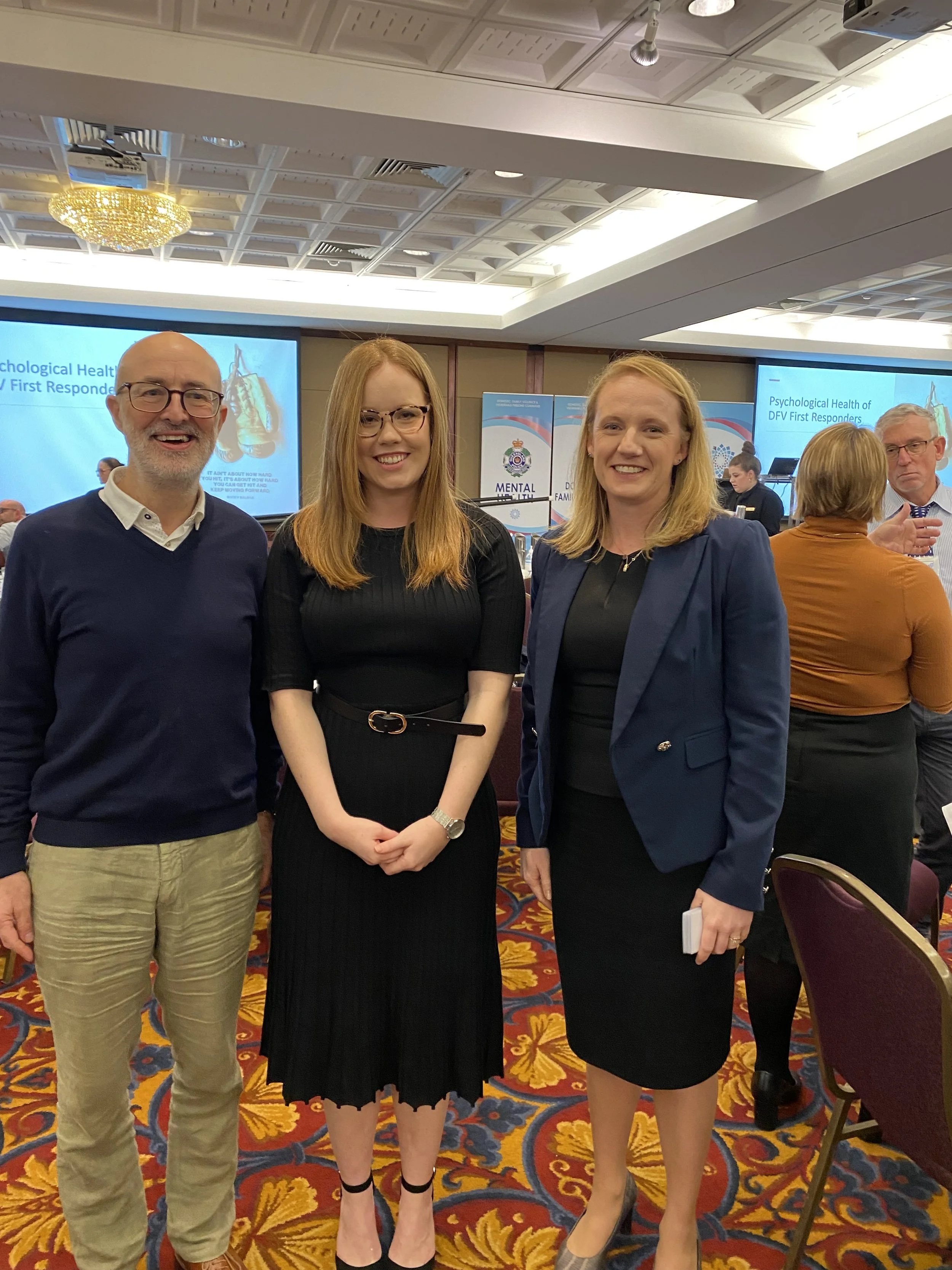 Group of three people smiling at a conference, with a presentation slide about psychological health of first responders in the background, and other attendees interacting in the event hall.