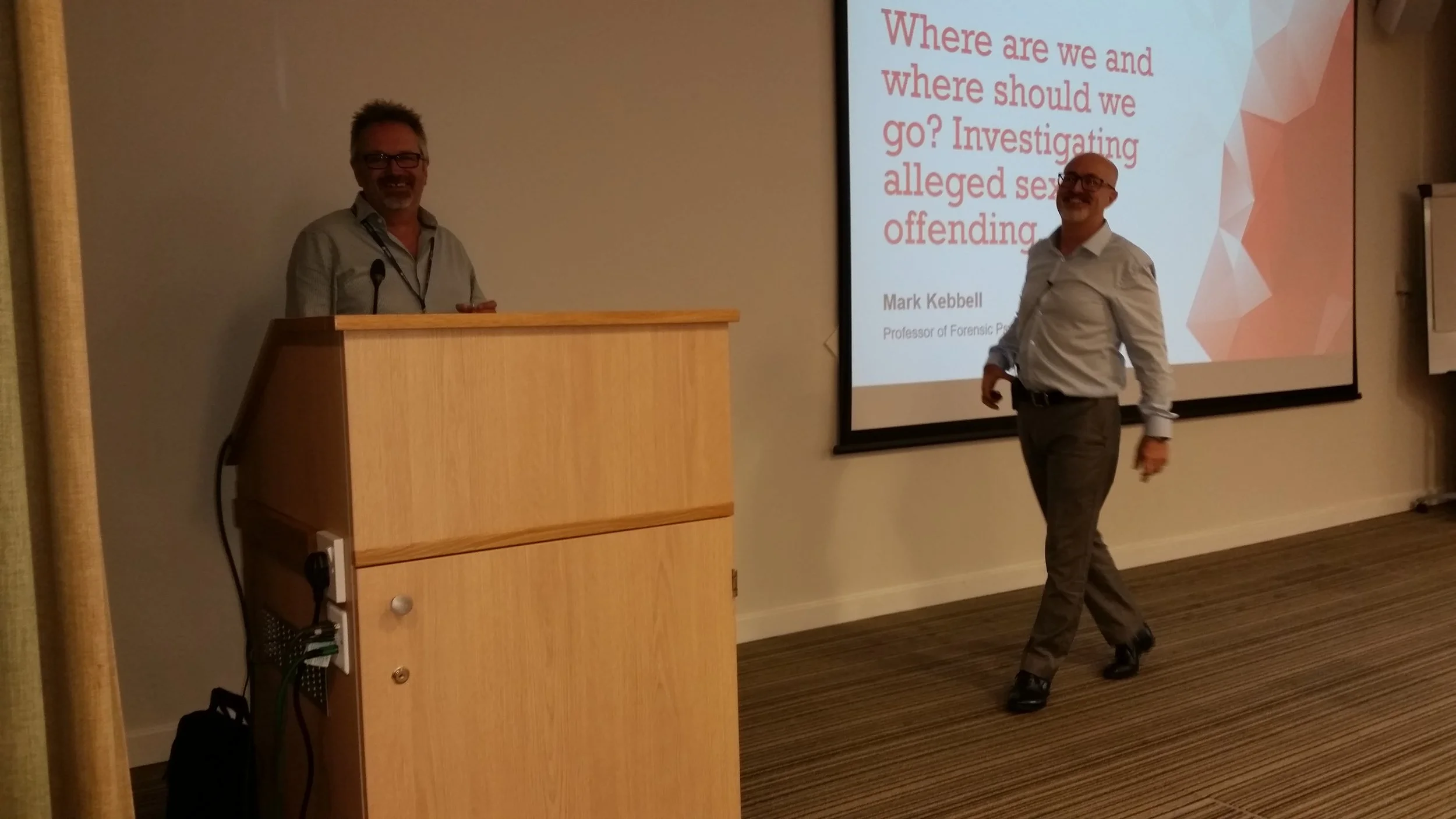 A man with glasses and a collared shirt standing at a podium with a microphone, another man with glasses wearing a white shirt walking past, and a presentation slide on the wall that reads 'Where are we and where should we go? Investigating alleged s