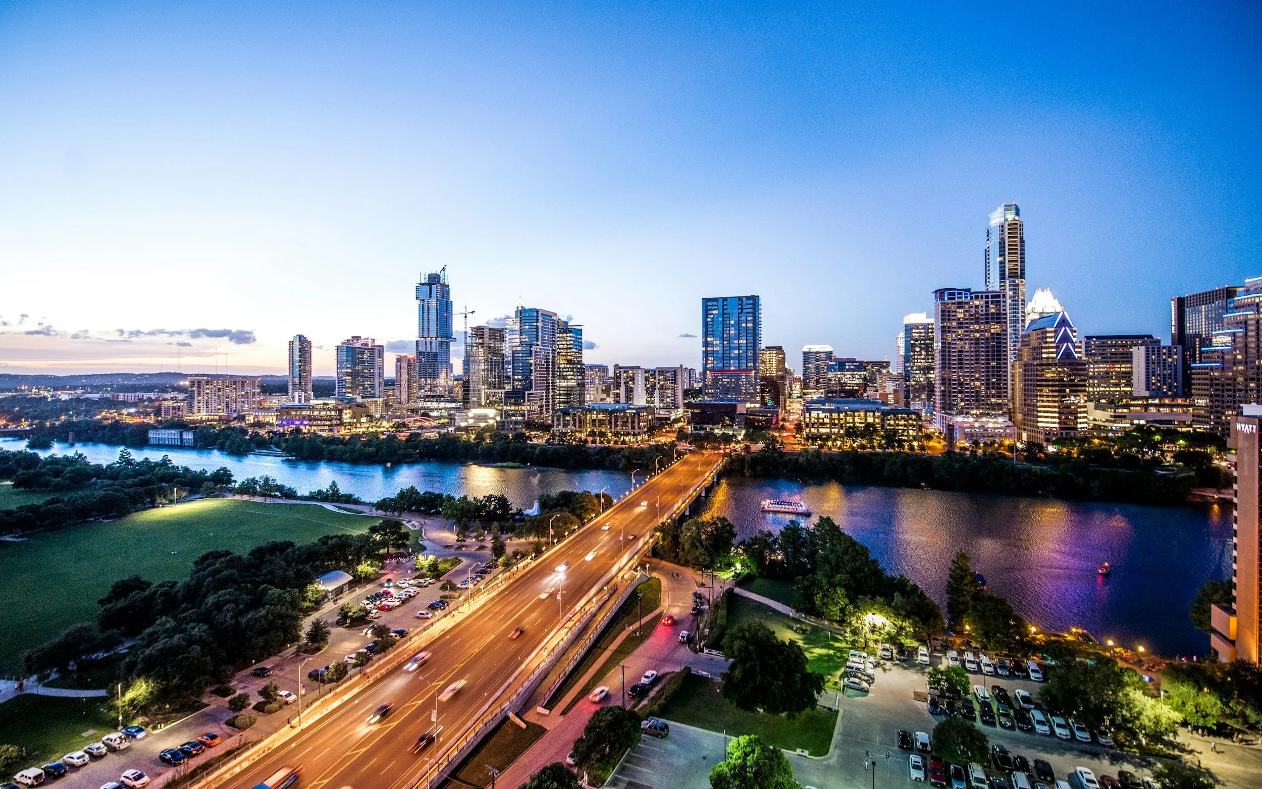 A city skyline at dusk with tall skyscrapers, a river, and a bridge crossing it. The sky is clear and the buildings are illuminated with lights.