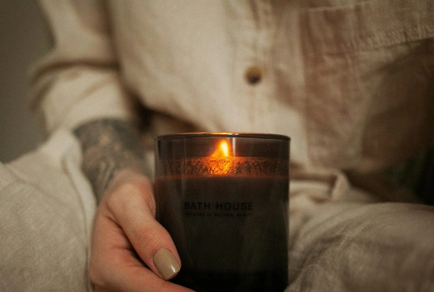 Person holding a lit Bath House scented candle with a dark glass container, casual clothing, and a tattooed arm, sitting in a cozy environment.