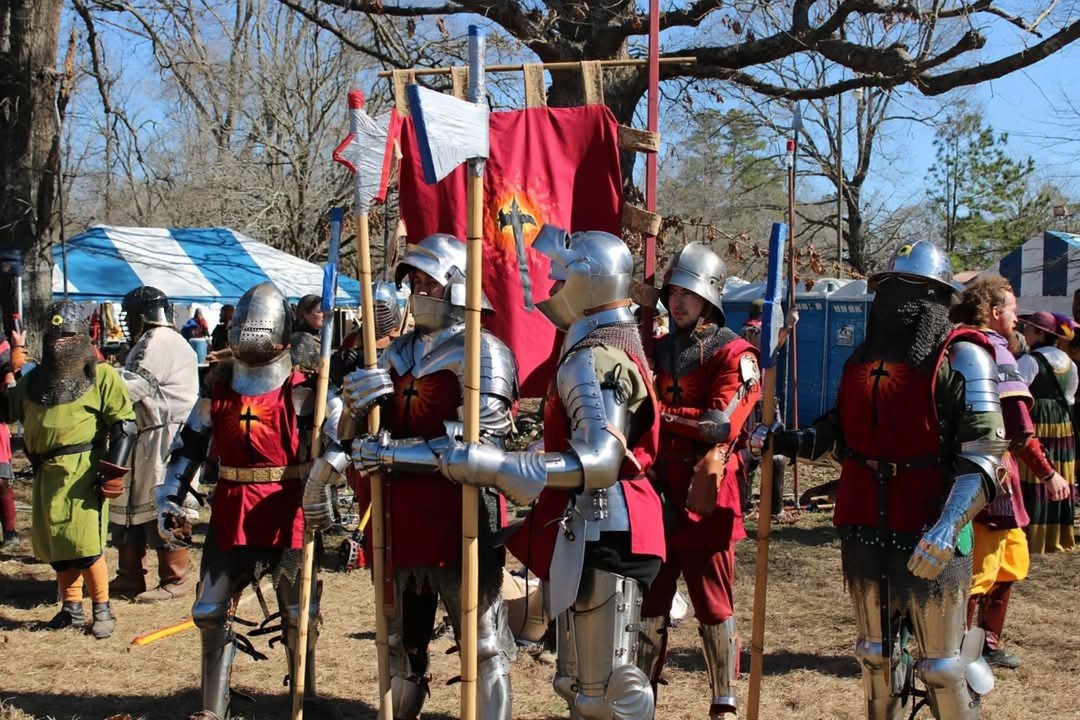 People dressed in medieval knight costumes with metallic armor, helmets, and shields gather outdoors with a red banner that has a flame symbol, under a large tree, during an event or festival.
