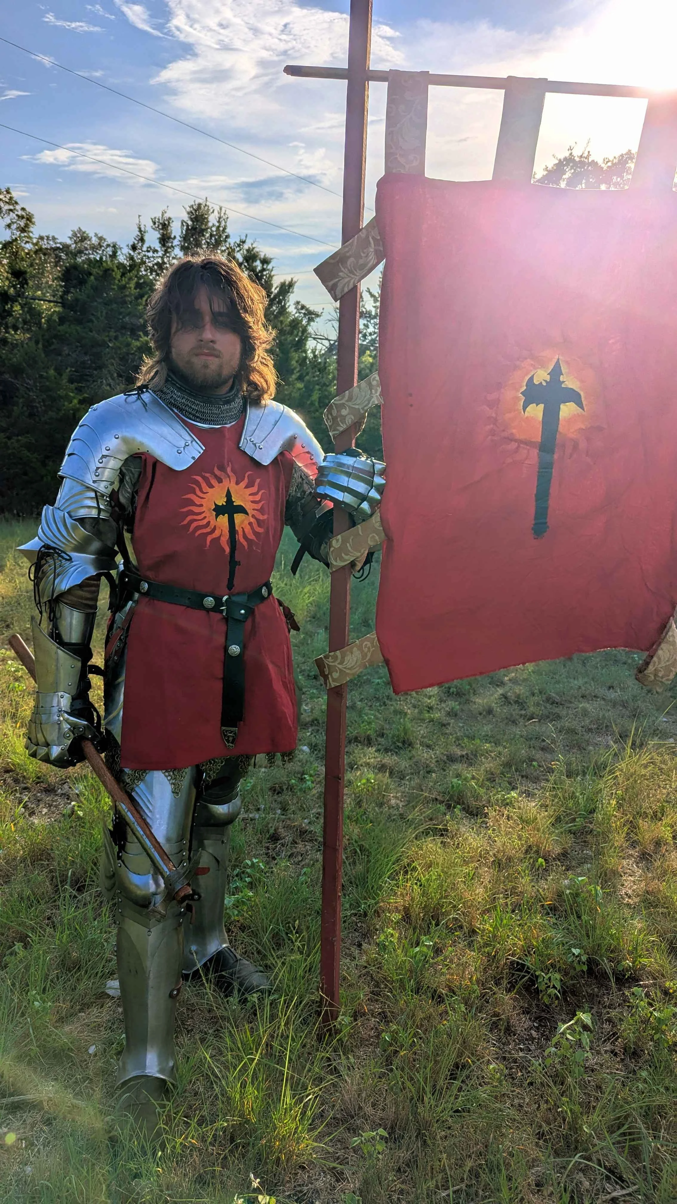 A person dressed as a medieval knight in armor and red tunic holding a flag with a dragon emblem, standing outdoors in a grassy area with trees and a blue sky.