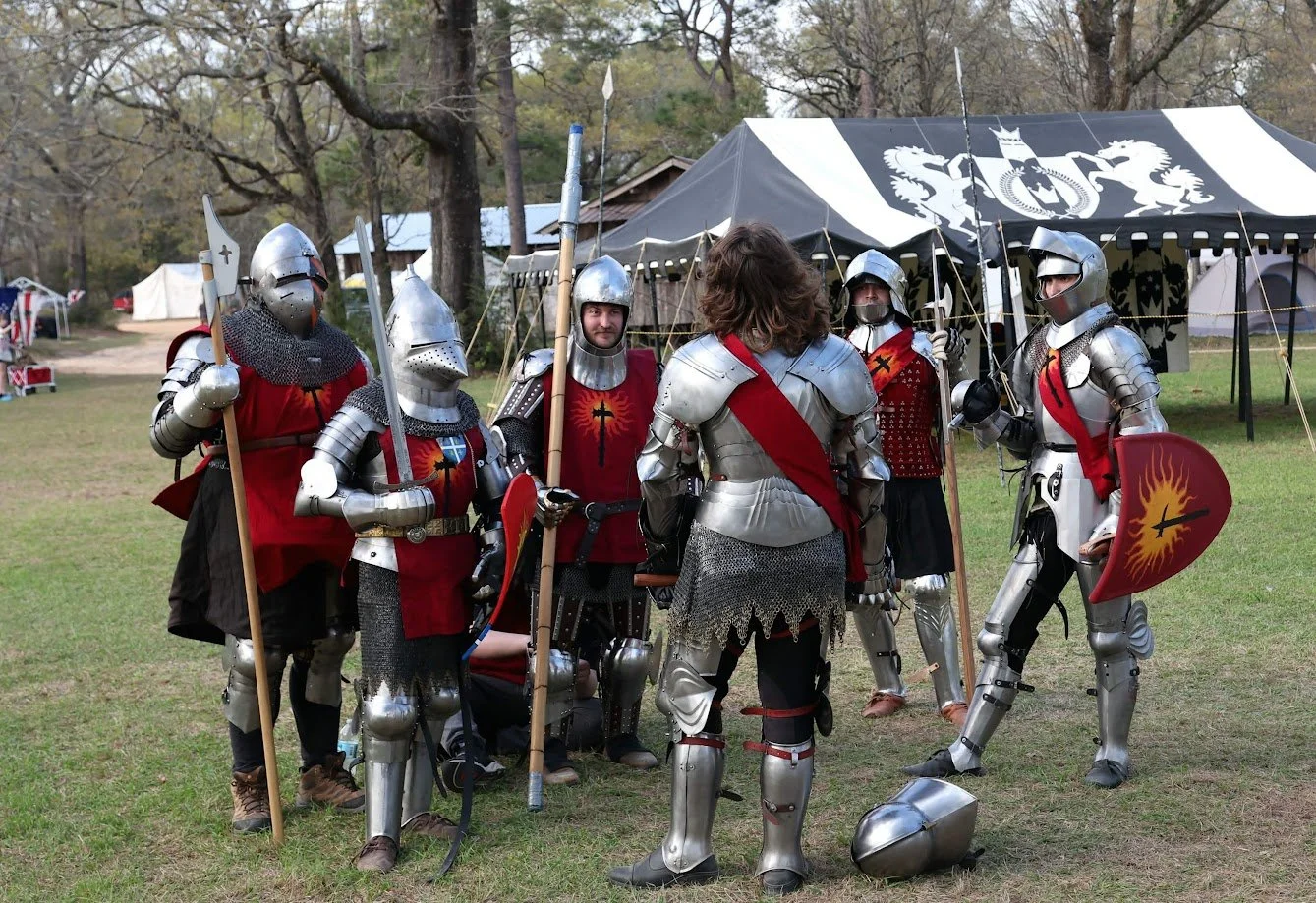 Group of people dressed in medieval knight costumes with armor, helmets, and shields gathered outdoors in front of a large tent with a black and white banner featuring heraldic lions. The scene appears to be a medieval reenactment or festival.