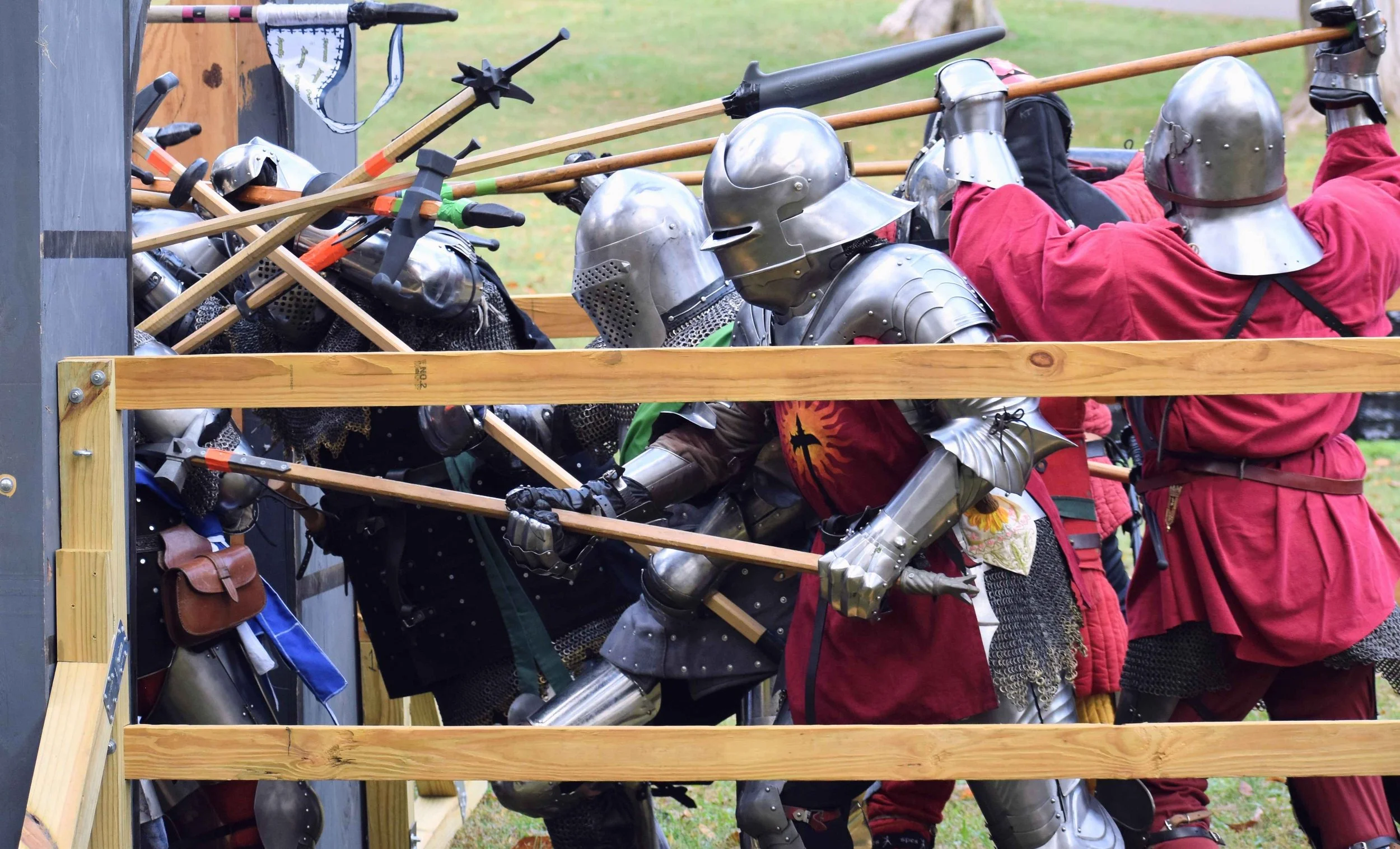 A group of people dressed in medieval armor and costumes, engaging in a mock battle or reenactment, with wooden barriers and outdoor setting.