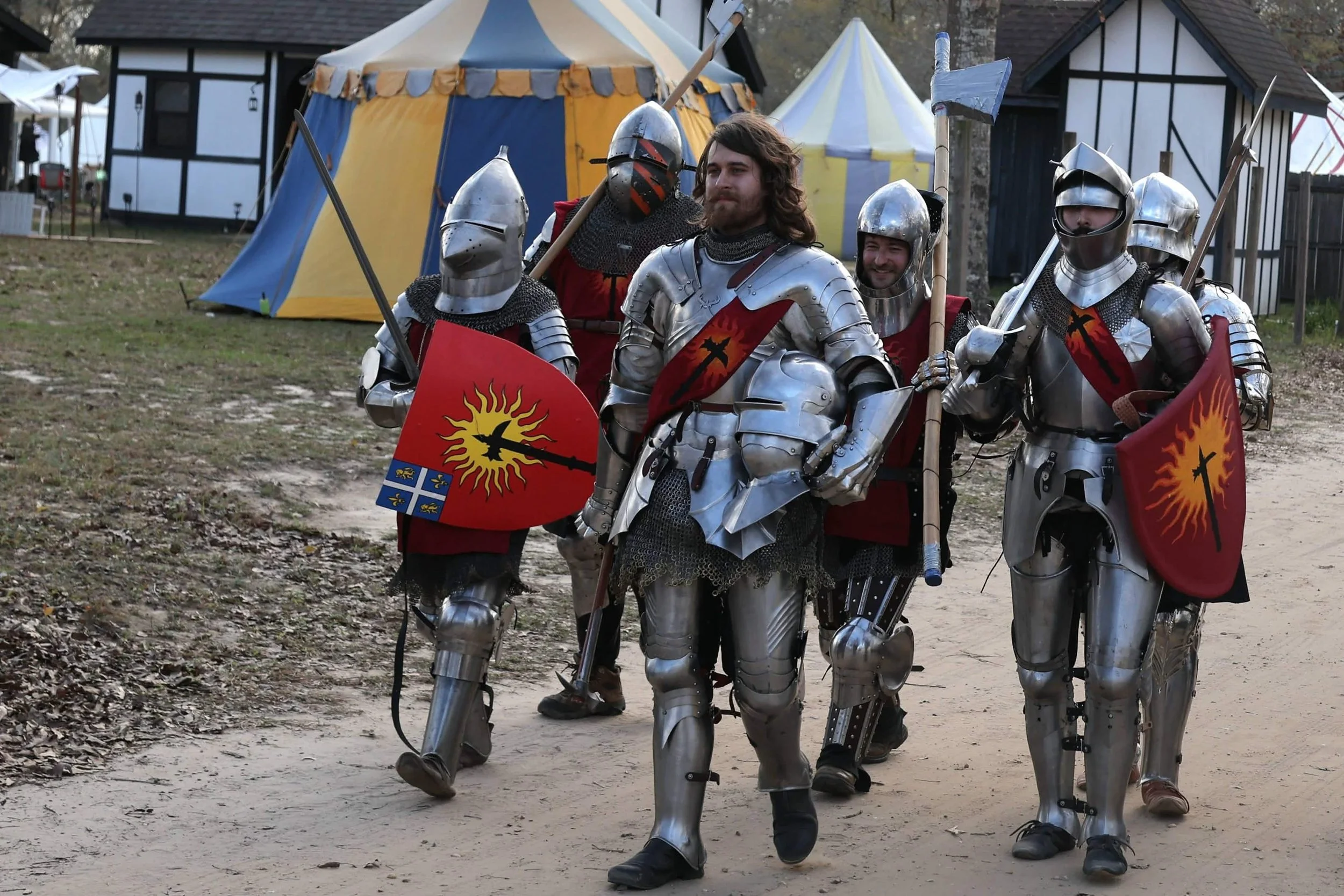 People dressed in medieval knight armor standing outdoors at a medieval fair, with colorful tents and buildings in the background.