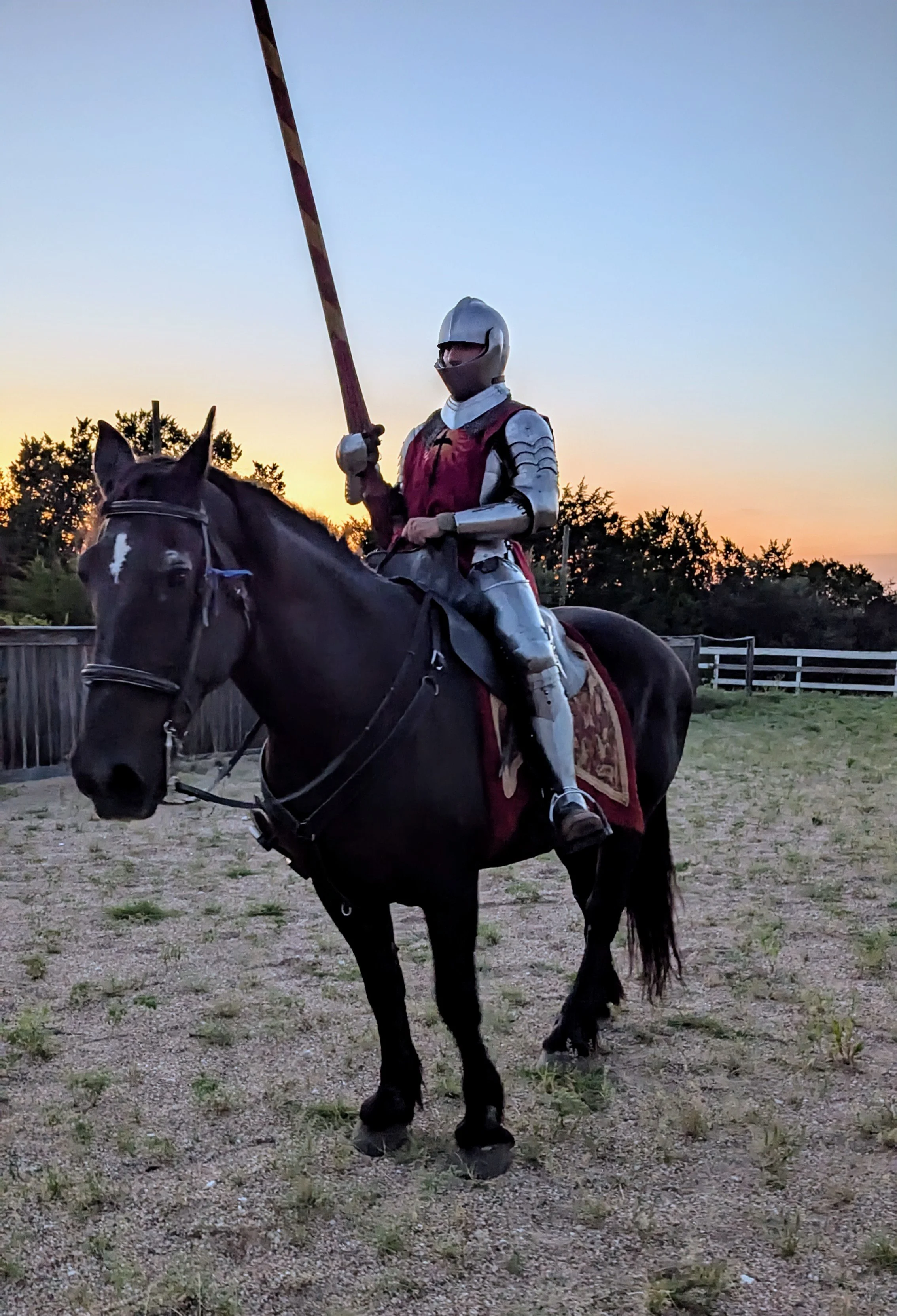 A knight in shiny armor riding a black horse with a saddle and original red and gold blanket, holding a pole with a flag at sunset, outdoors with trees and a wooden fence in the background.