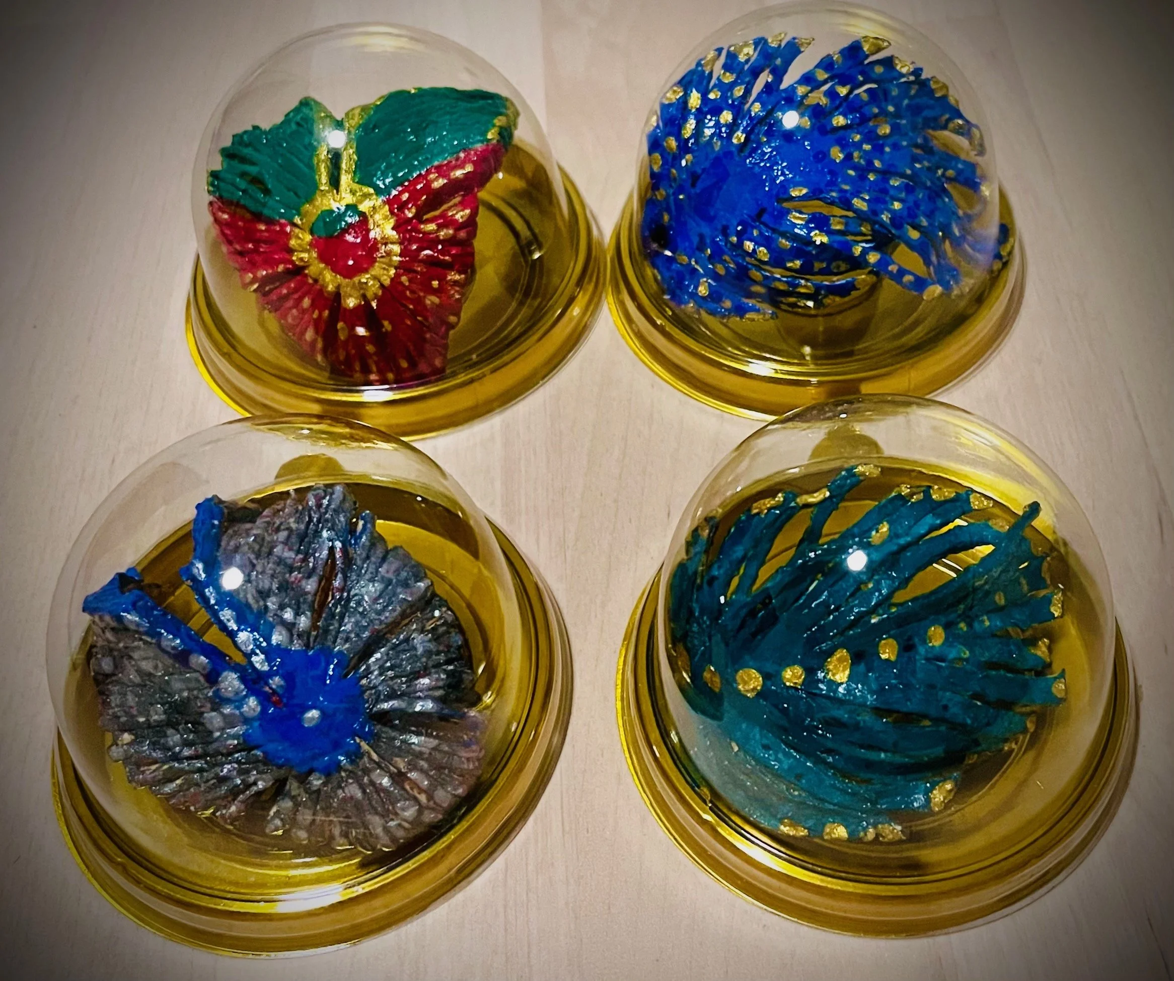 Colorful designer citrus rind ornaments in clear plastic containers with gold bases, featuring intricate, swirling, and patterned decorations in bright colors like red, green, blue, and silver.
