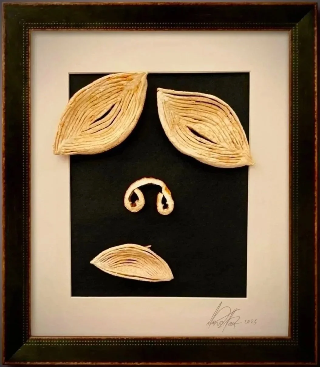Framed artwork made of layered dried citrus rinds resembling a human face, with two large leaf-like shapes for eyebrows, a small curled shape for a nose, and a leaf for a mouth.