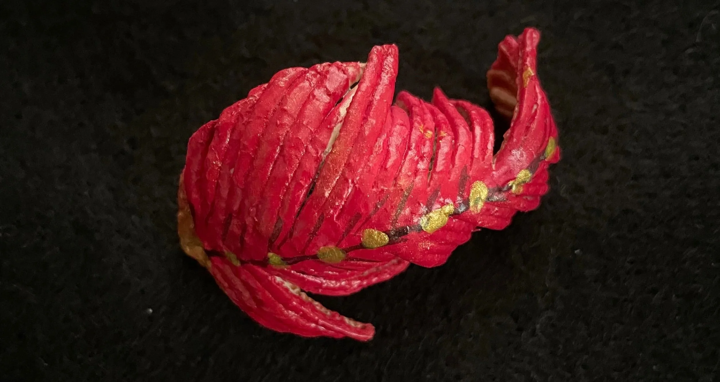 A red flower made from a citrus rind with gold accents, placed on a black textured surface.