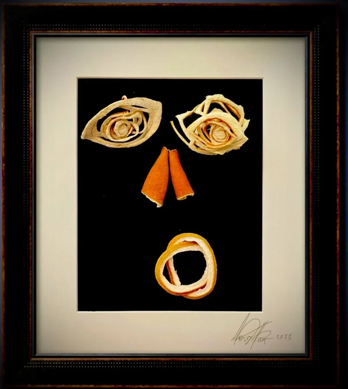 Artistic face made from dried citrus rinds arranged on a black background, framed in a decorative black frame.