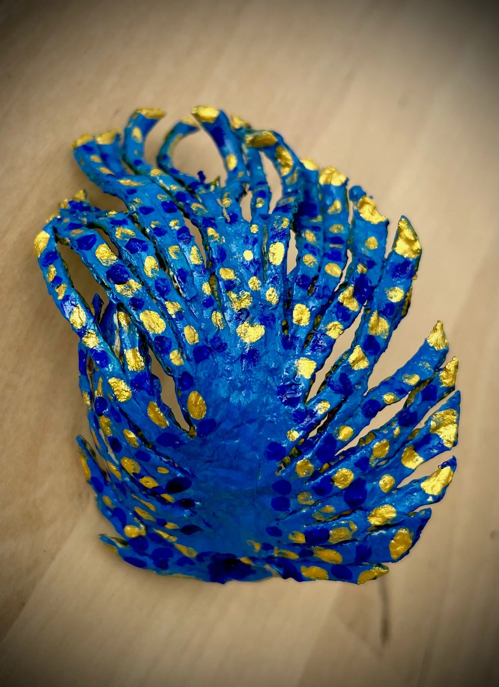 Blue citrus rind sculpture with gold dots, shaped like a flower or sea creature, placed on a beige surface.