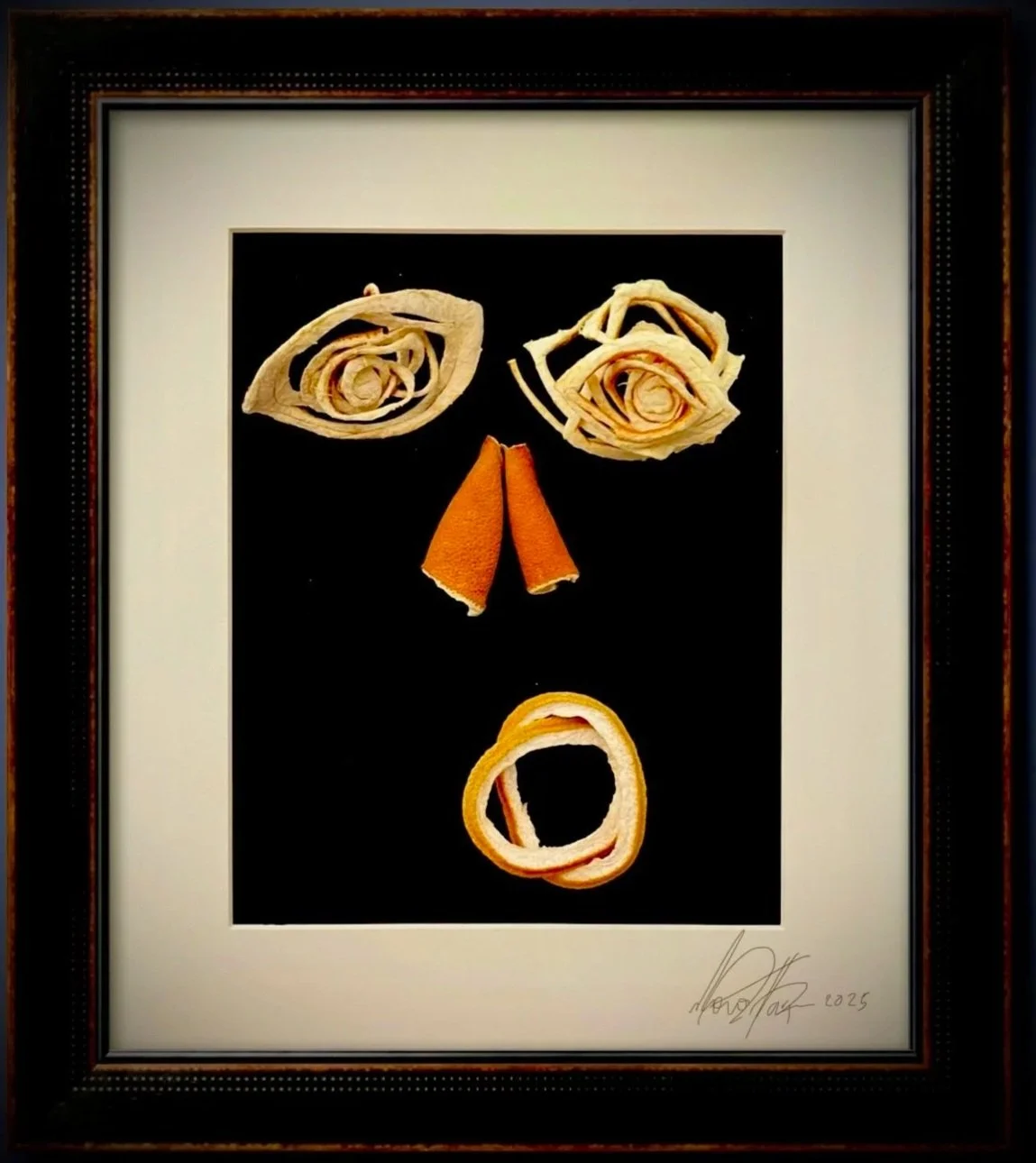 Framed artwork of various curled and folded orange and beige citrus rinds arranged on a black background, resembling a face with eyes, nose, and mouth.