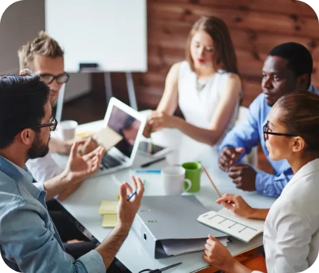 Group of six digital marketing experts in a meeting room having a discussion, with laptops, notebooks, and coffee cups on the table.