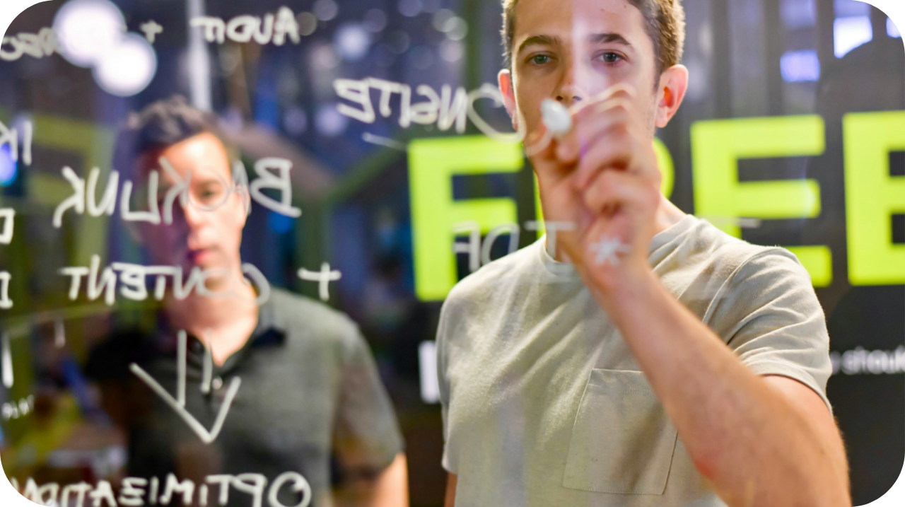 Two men writing on a glass wall with a marker, one in focus and the other blurred in the background.