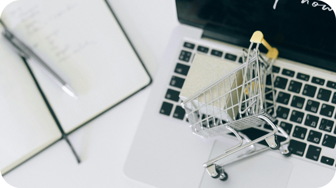 Close-up of a miniature shopping cart filled with sticky notes placed on a laptop keyboard, with a notebook and pen nearby on a white surface.