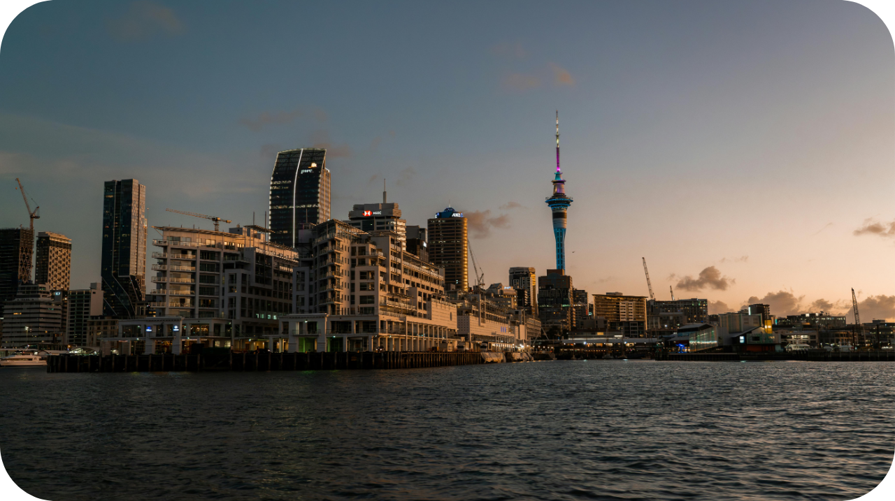 City skyline at dusk showing tall buildings, construction cranes, and a prominent tower with colorful lights, viewed across a body of water.