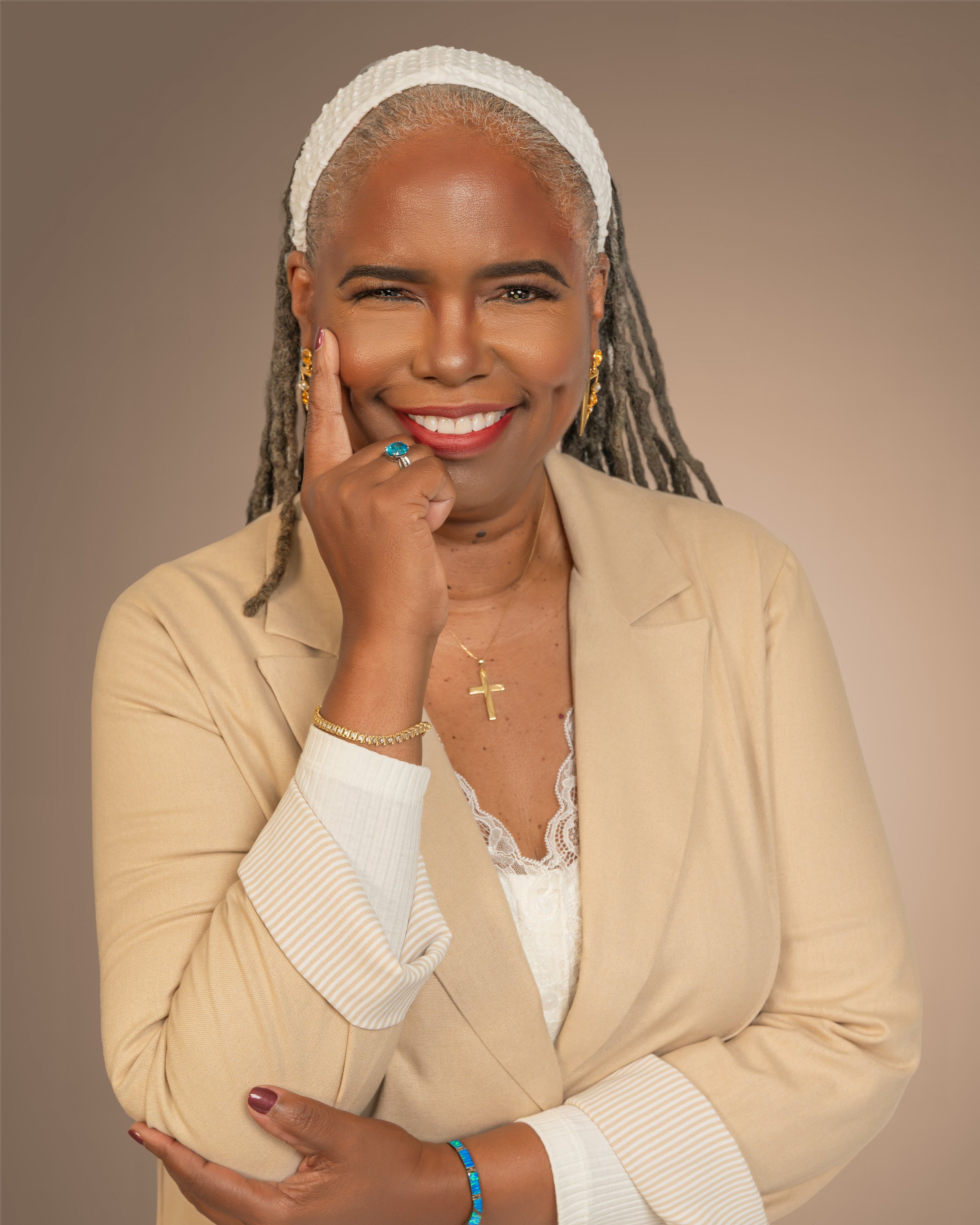 A smiling woman with gray dreadlocks, wearing a white headband, gold earrings, a gold cross necklace, a blue beaded bracelet, and a light beige blazer, standing against a neutral background.