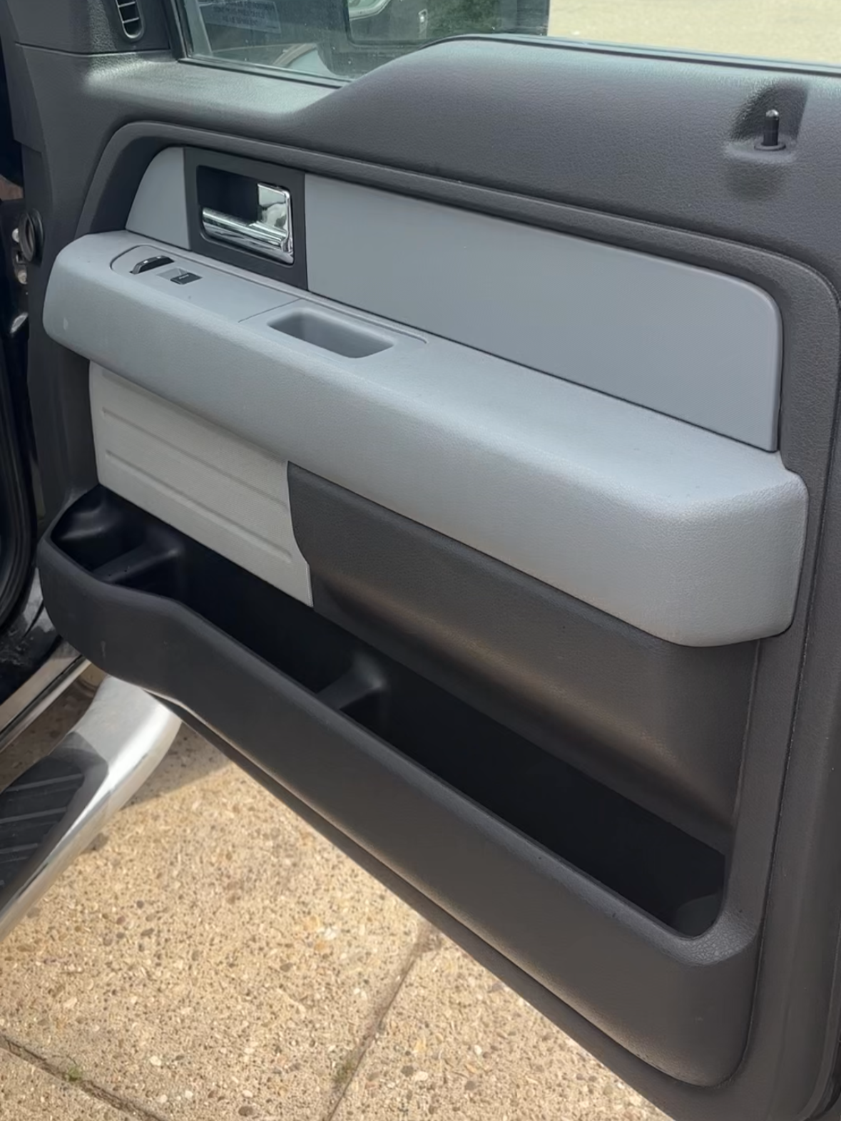 The interior of a vehicle's driver side door, featuring a door handle, window controls, and a storage compartment, with a view of the outside pavement and part of a vehicle in the background.