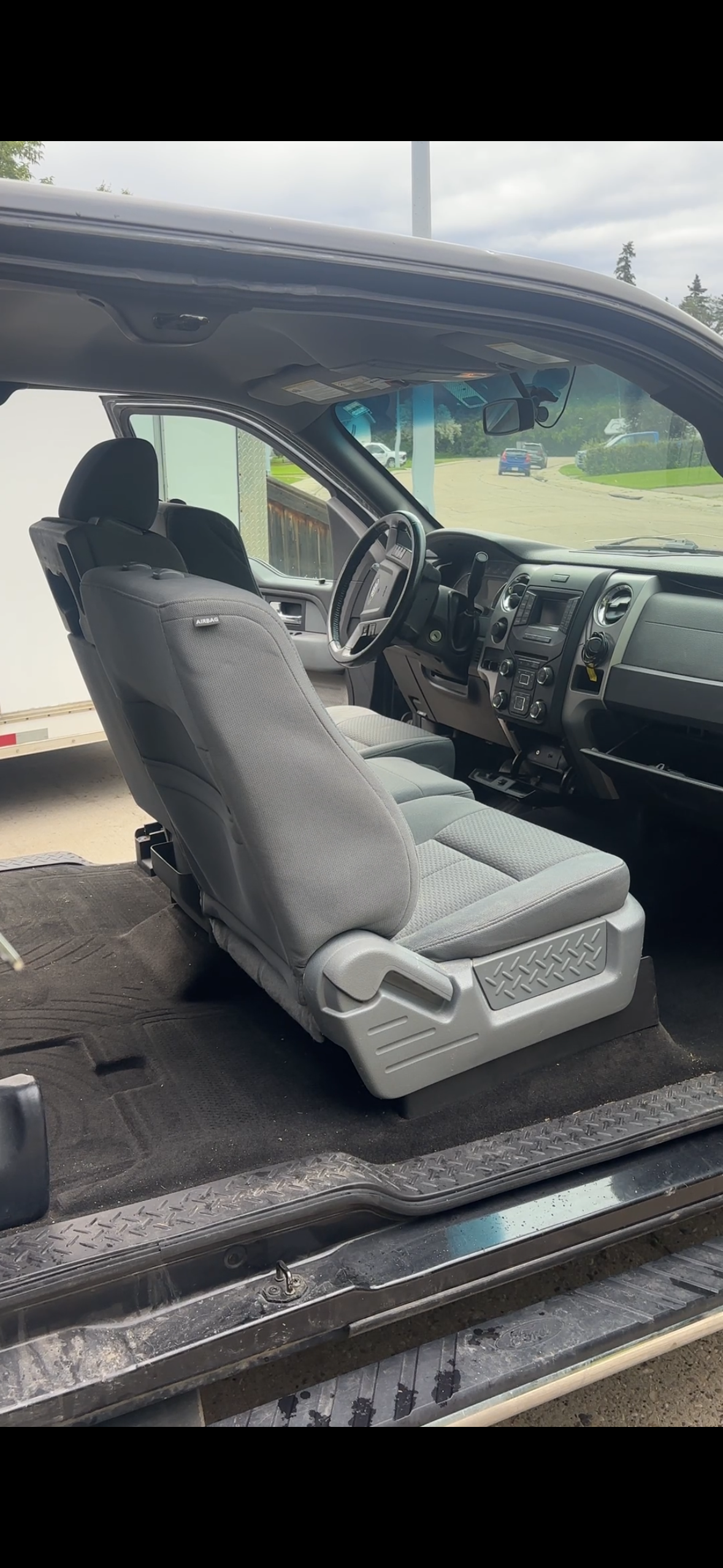 Inside of a vehicle showing the driver's and passenger's seats, with a view of the dashboard and windshield. The seats are gray, and the floor is carpeted with black mats. Outside the vehicle, there are parked cars and a cloudy sky.