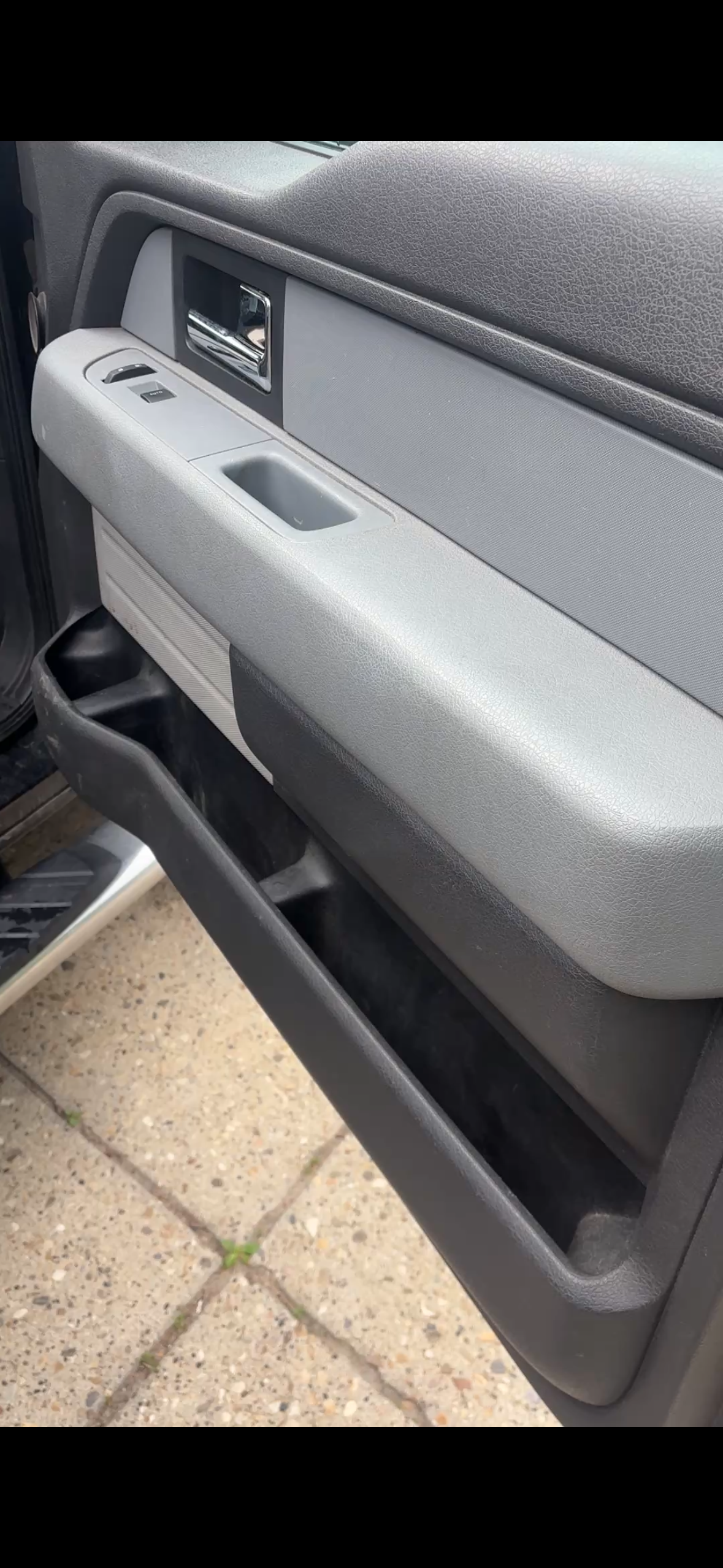 Inside of a vehicle door showing a grey armrest, door handle, window controls, and a black storage compartment near the floor.