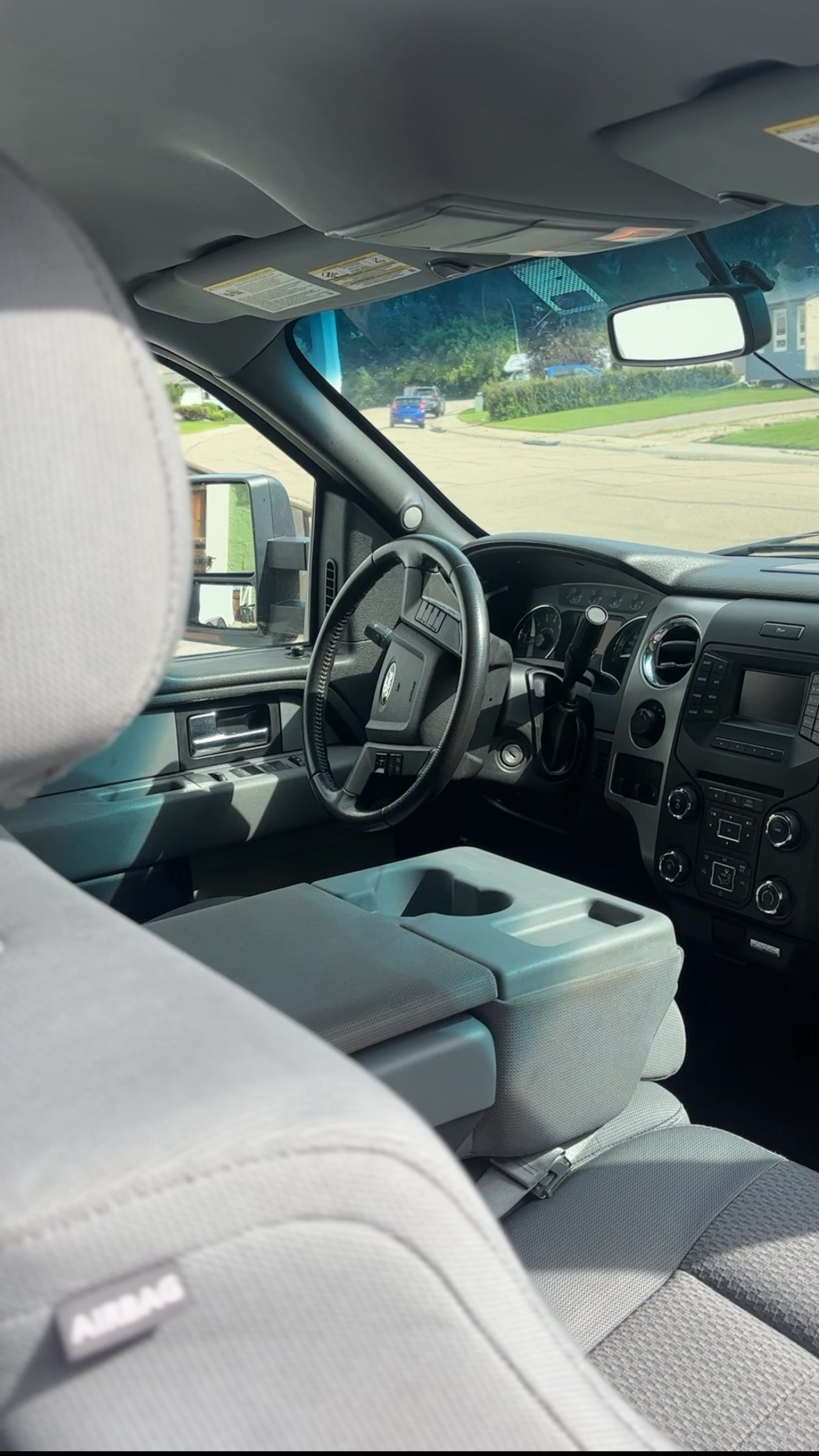 Inside view of a vehicle interior showing front seats, dashboard with controls, steering wheel, rearview mirror, and windshield with a view of the street outside.