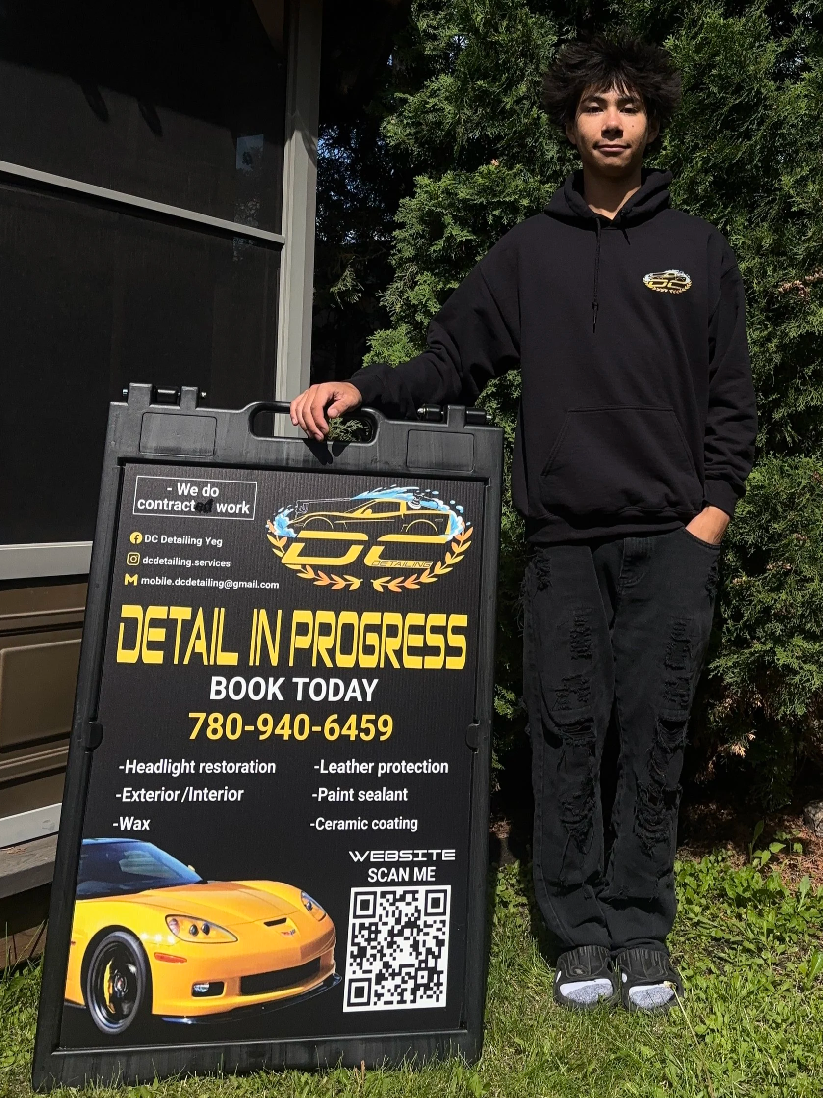 A man standing outdoors next to a blackboard sign for a detailing service, with a background of green shrubs and trees. The sign features a yellow sports car, contact information, a QR code, and list of services.