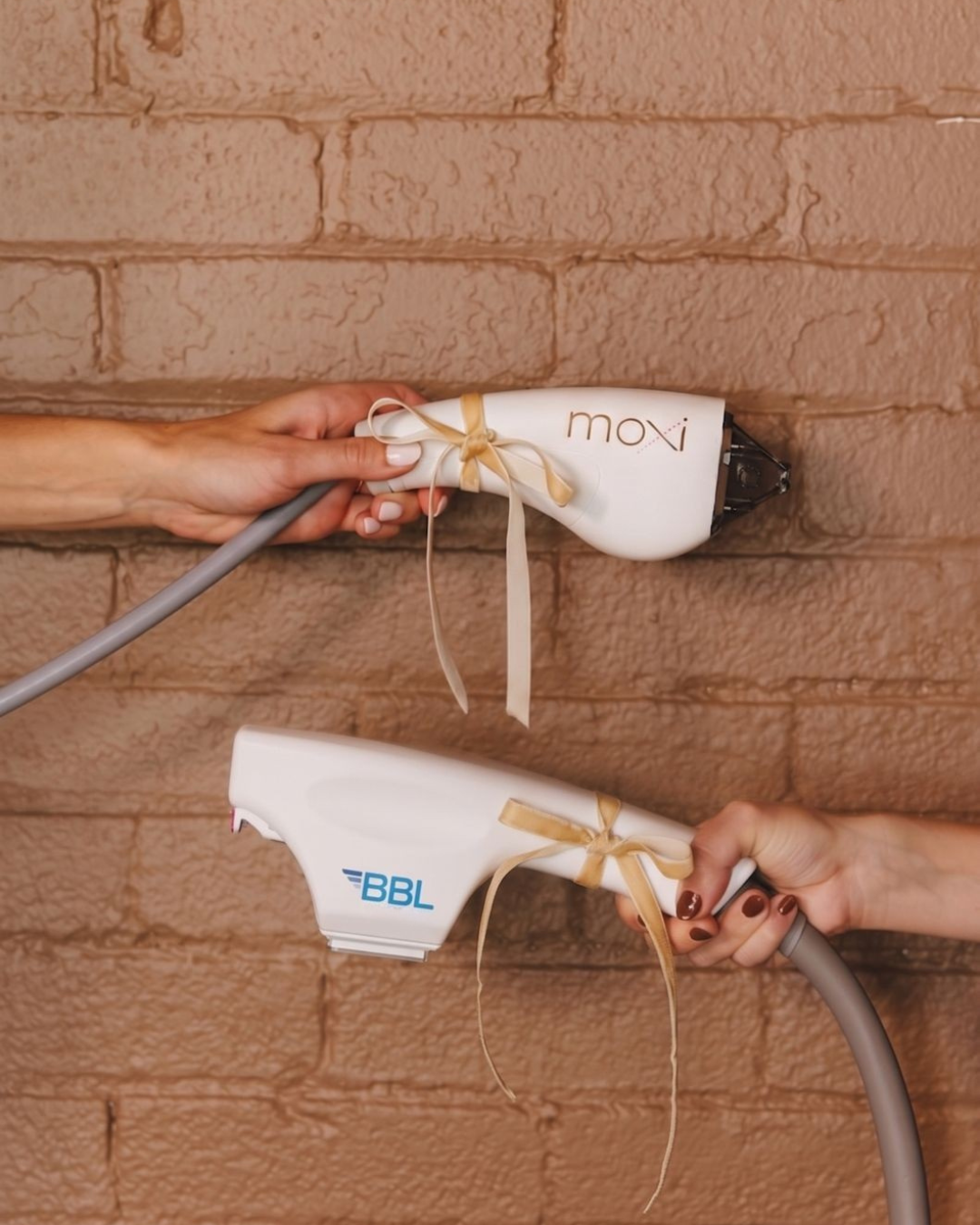 Two hands holding hairdryers, one labeled 'moxi' and the other labeled 'BBL', against a brick wall, with ribbons tied around the handles.