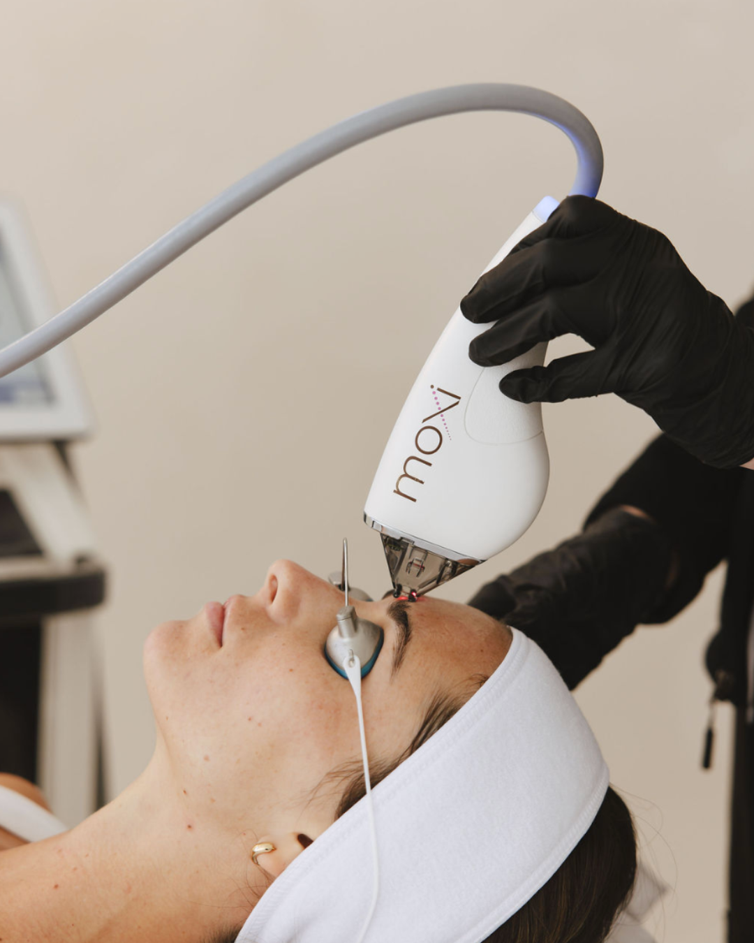 A person receiving a laser facial treatment from a professional wearing black gloves, using a white device labeled 'Vow' to treat the person's forehead while the person lies on a treatment bed with a white eye mask and headband.