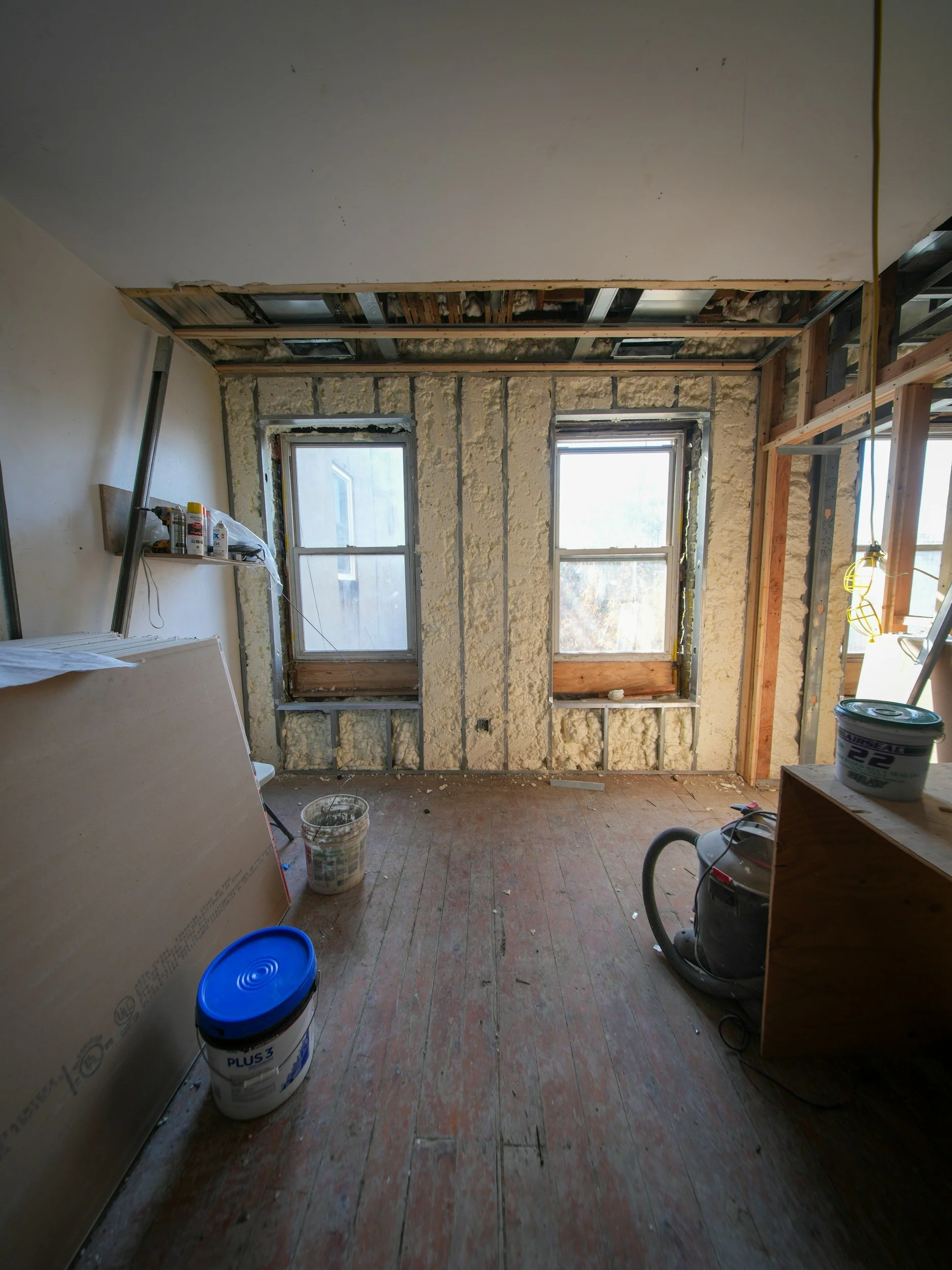 Interior of a room under renovation showing exposed walls and ceiling with insulation, two windows, construction tools and materials, including a vacuum cleaner, buckets, cans of paint, and wooden framing.