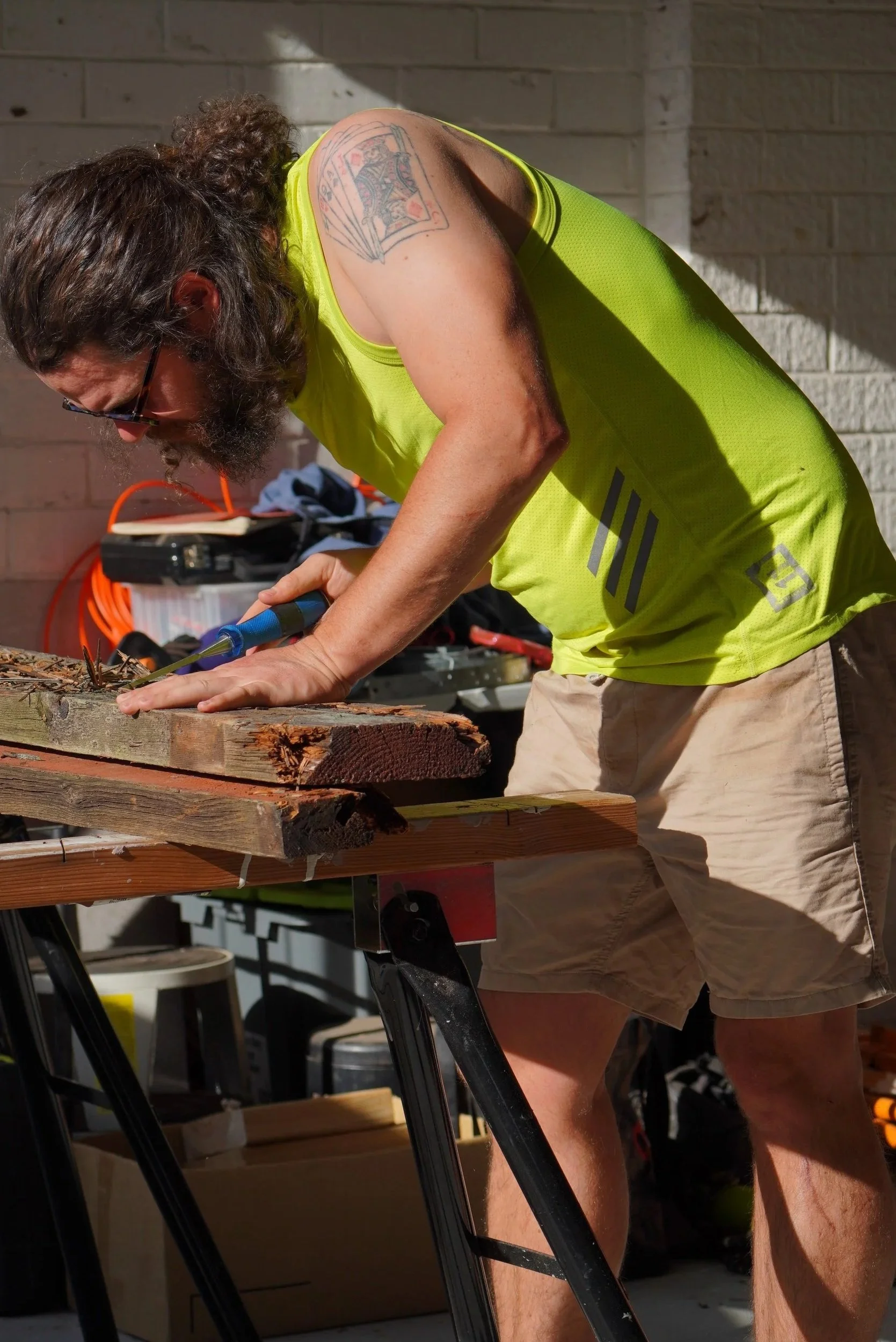 A man with long curly hair and a beard wearing sunglasses, a bright yellow tank top, and beige shorts is working on a woodworking project using a hand tool on a piece of wood in a workshop or garage.