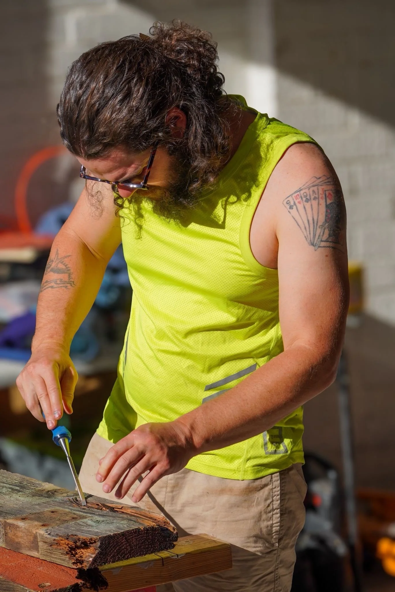 A man with long curly hair and tattoos on his arm working on a piece of wood using a screwdriver or chisel, wearing a bright yellow sleeveless shirt in a workshop.